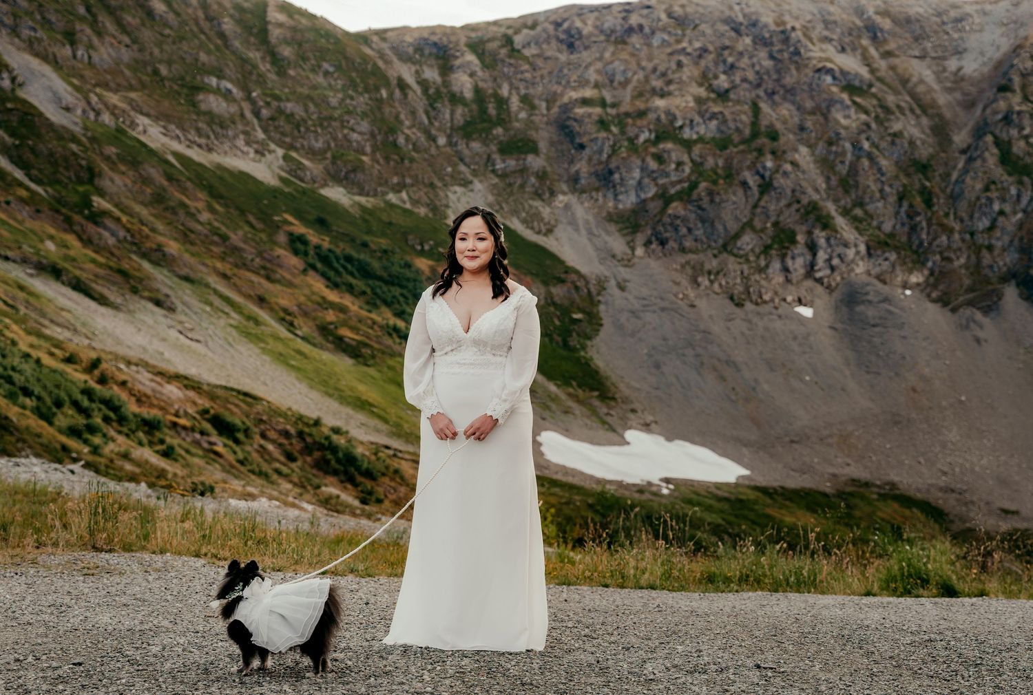 Bride in white gown poses with black and white dog on mountain landscape with alpine lake backdrop.