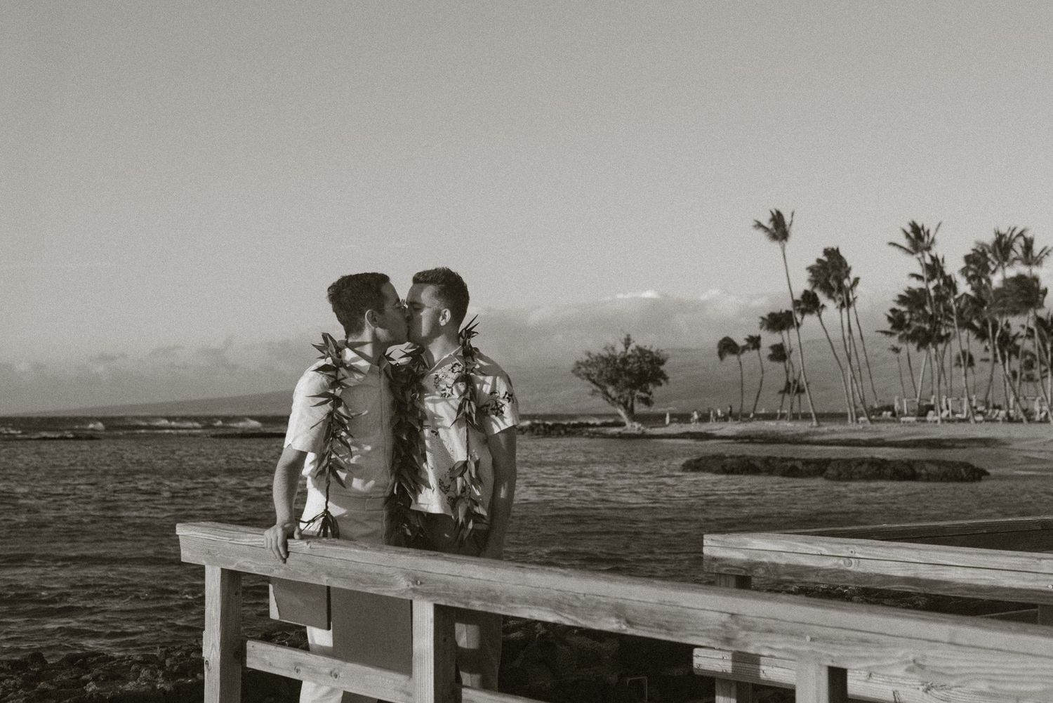Couple sharing a romantic kiss on a wooden pier overlooking a tropical beach with palm trees in black and white.