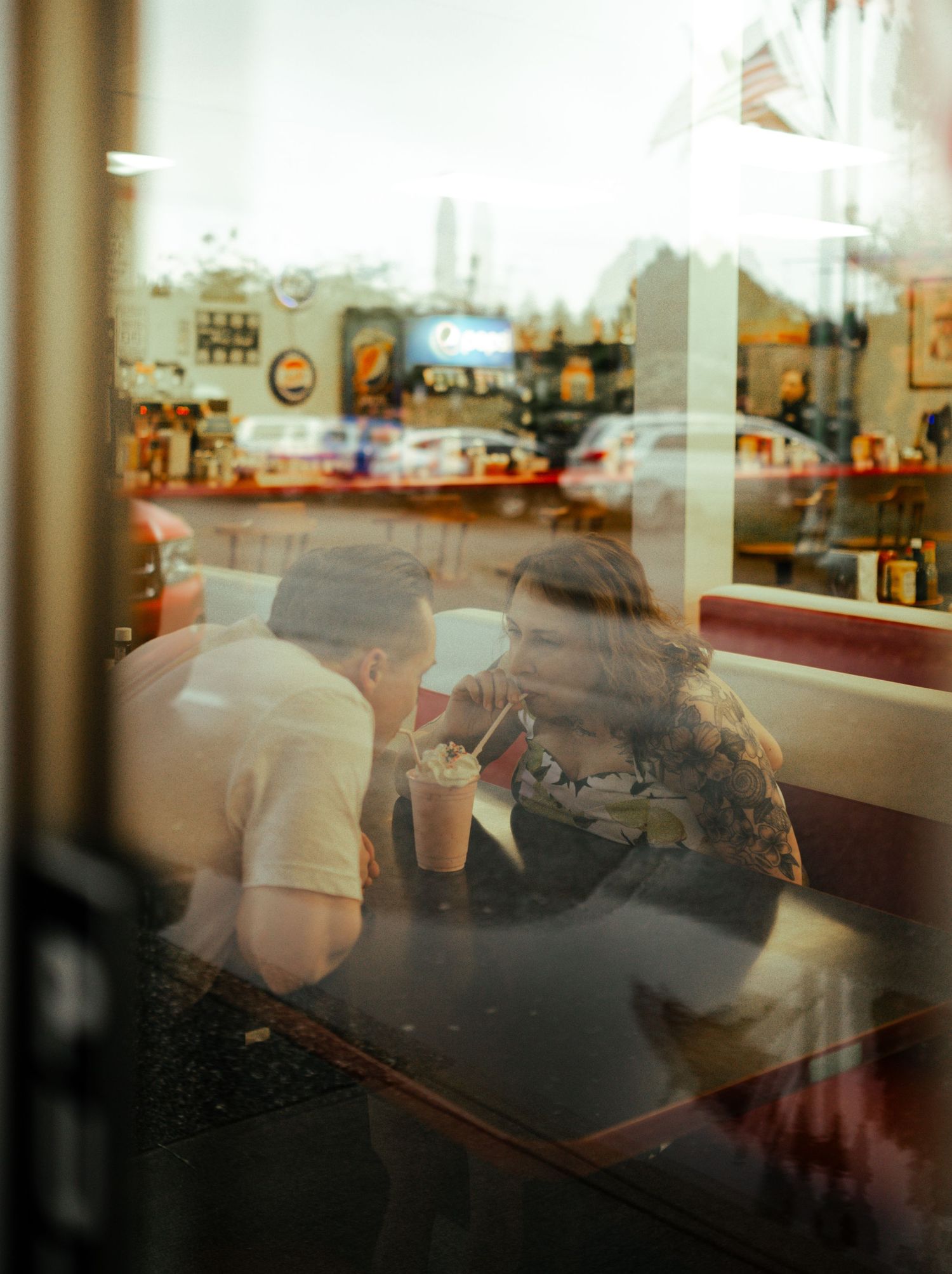 Two people sharing a milkshake at a retro diner booth, photographed through a window reflection in cafe in Ketchikan Alaska