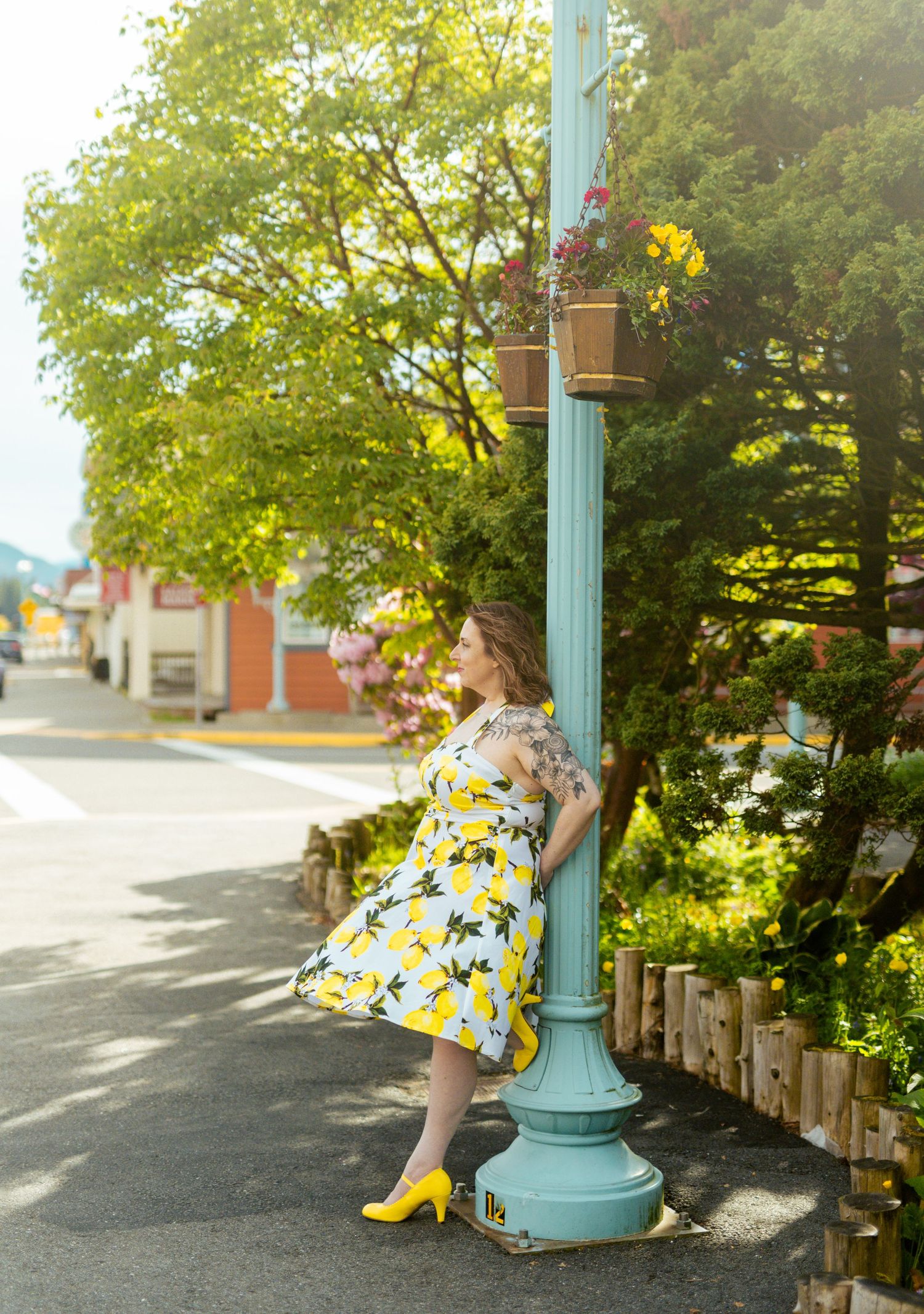 Woman in yellow lemon-print dress and heels leaning against a teal lamp post on a sunny small-town street in Ketchikan Alaska during Summer