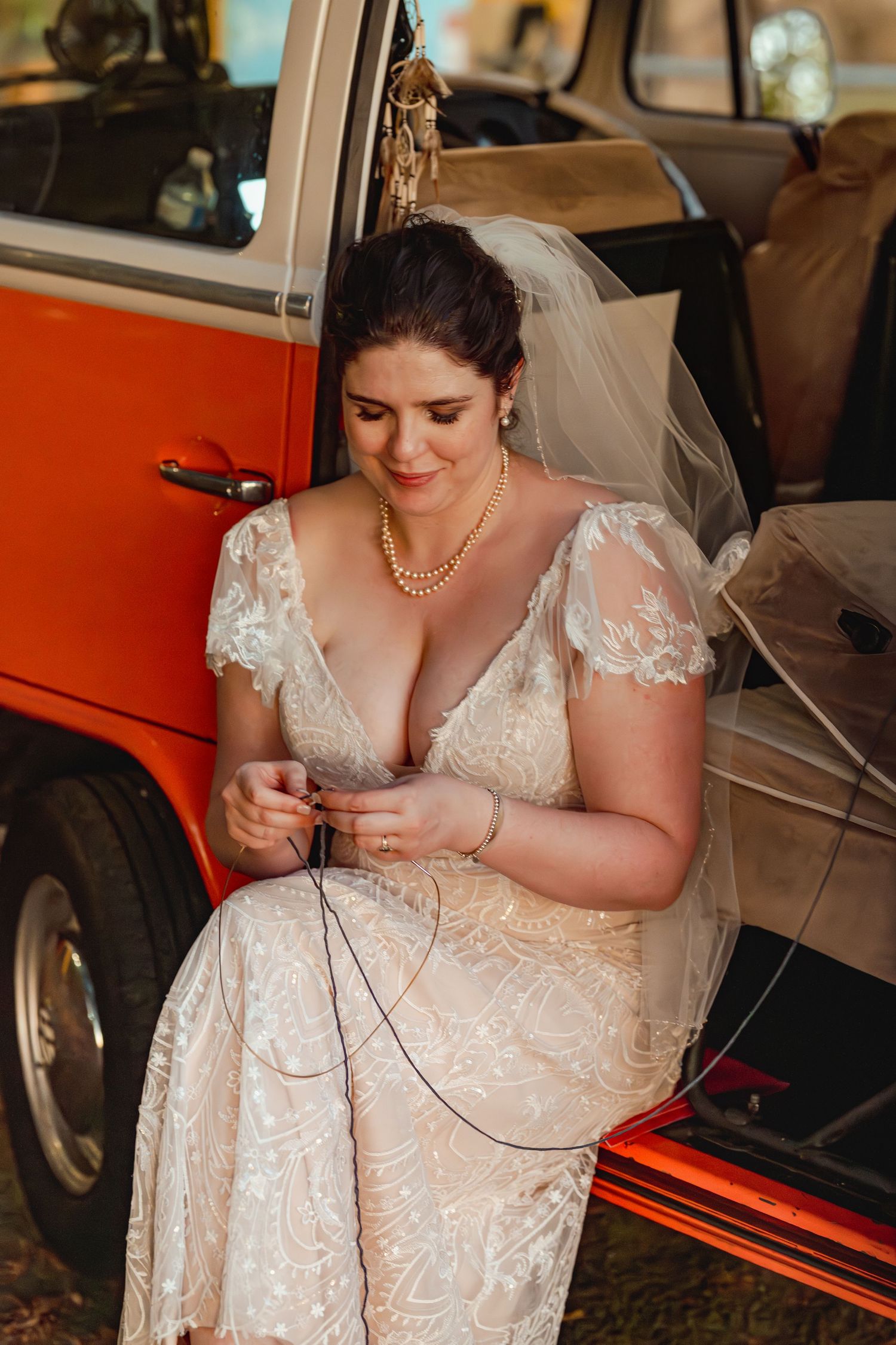 Bride in lace wedding dress and veil sits in vintage orange VW van, adjusting jewelry with pearl necklace.