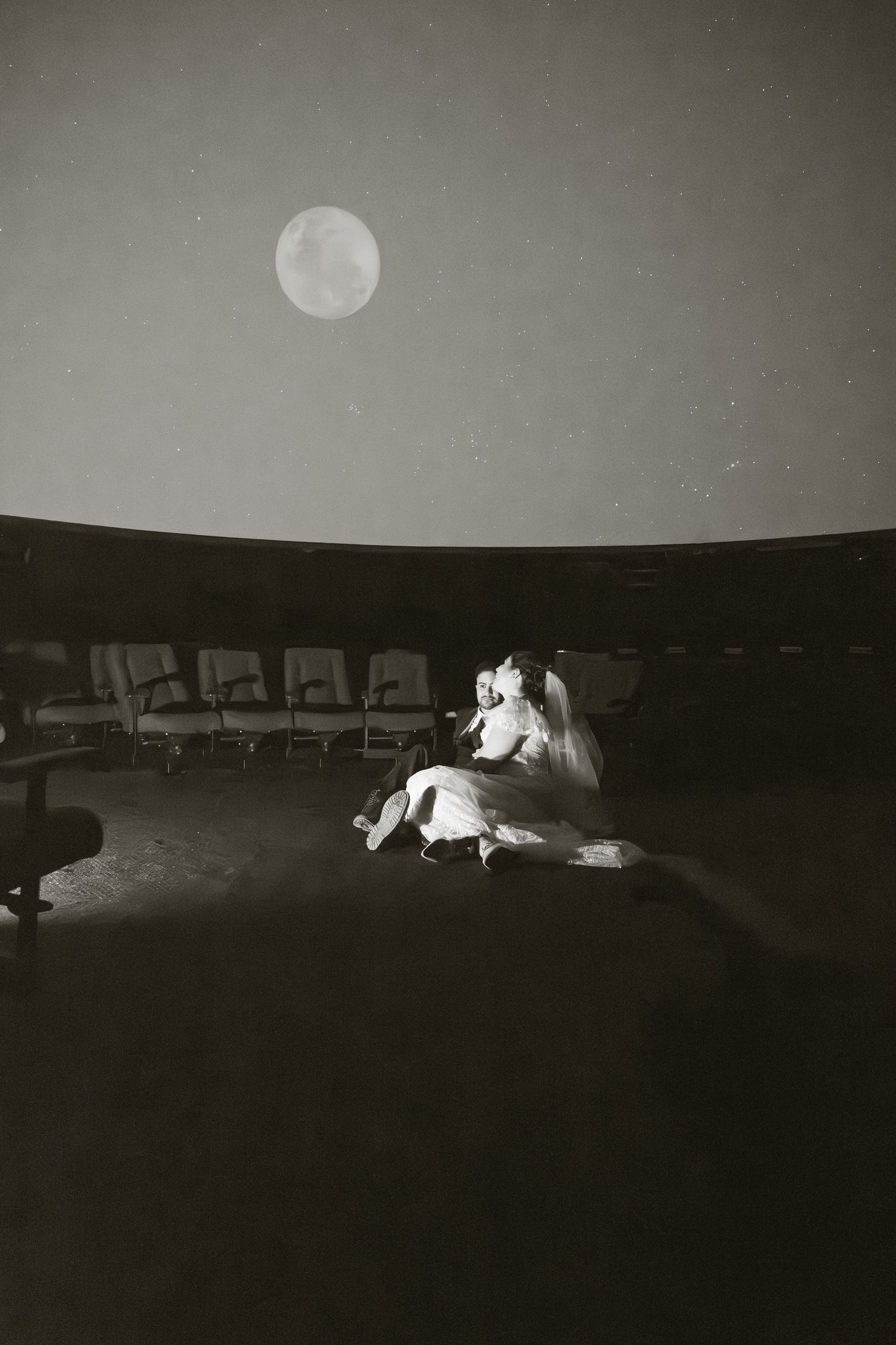 Bride and groom sitting under a glowing moon inside a planetarium in a romantic black and white photo.