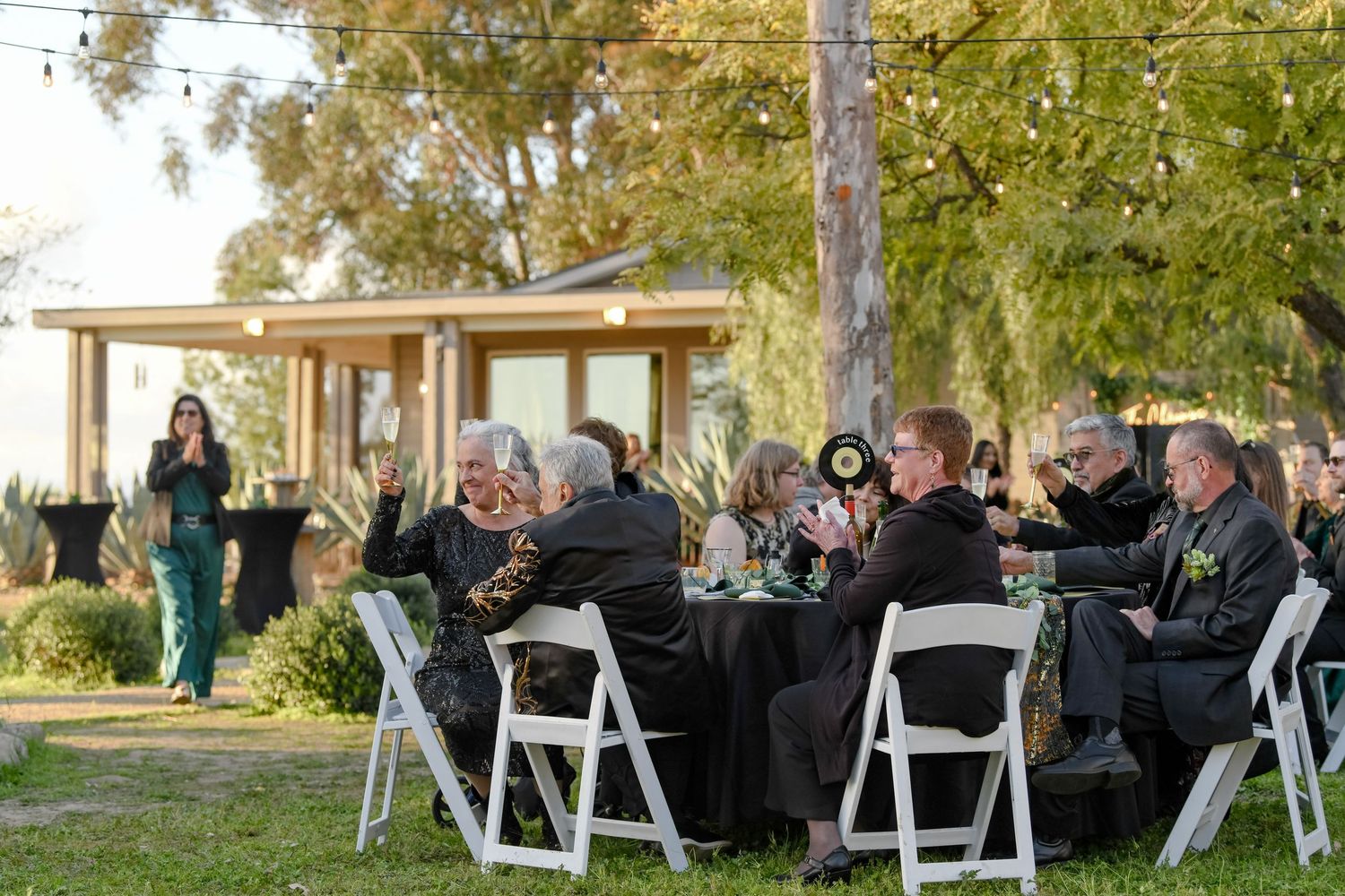 Wedding guests toasting and enjoying a casual outdoor reception with white folding chairs and black tablecloths.