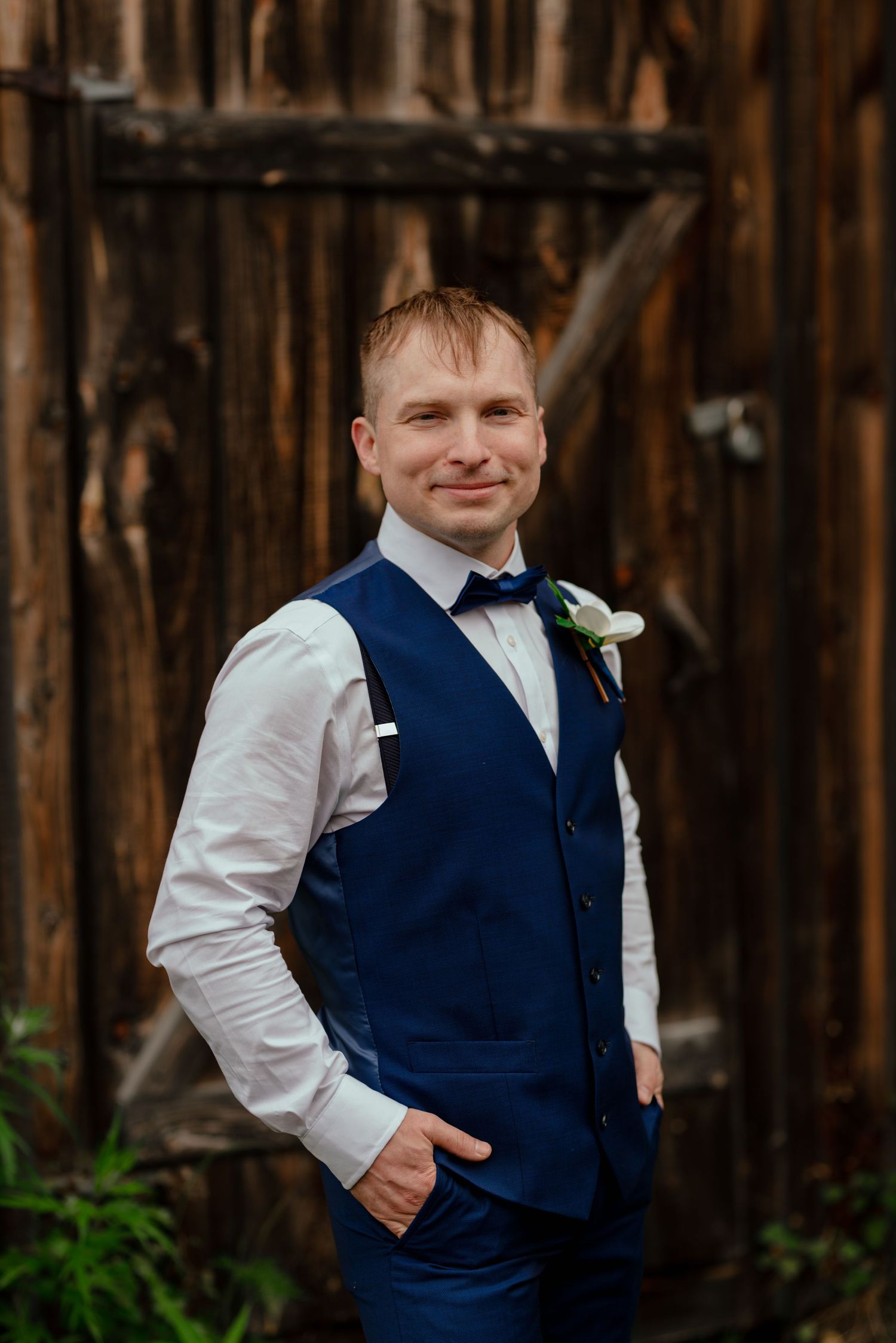Groom in navy blue vest and bow tie with boutonniere poses confidently before rustic wooden barn doors.