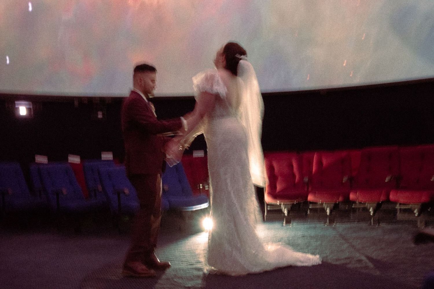 A couple exchanges wedding vows in a stunning planetarium venue with red seats and a domed ceiling.