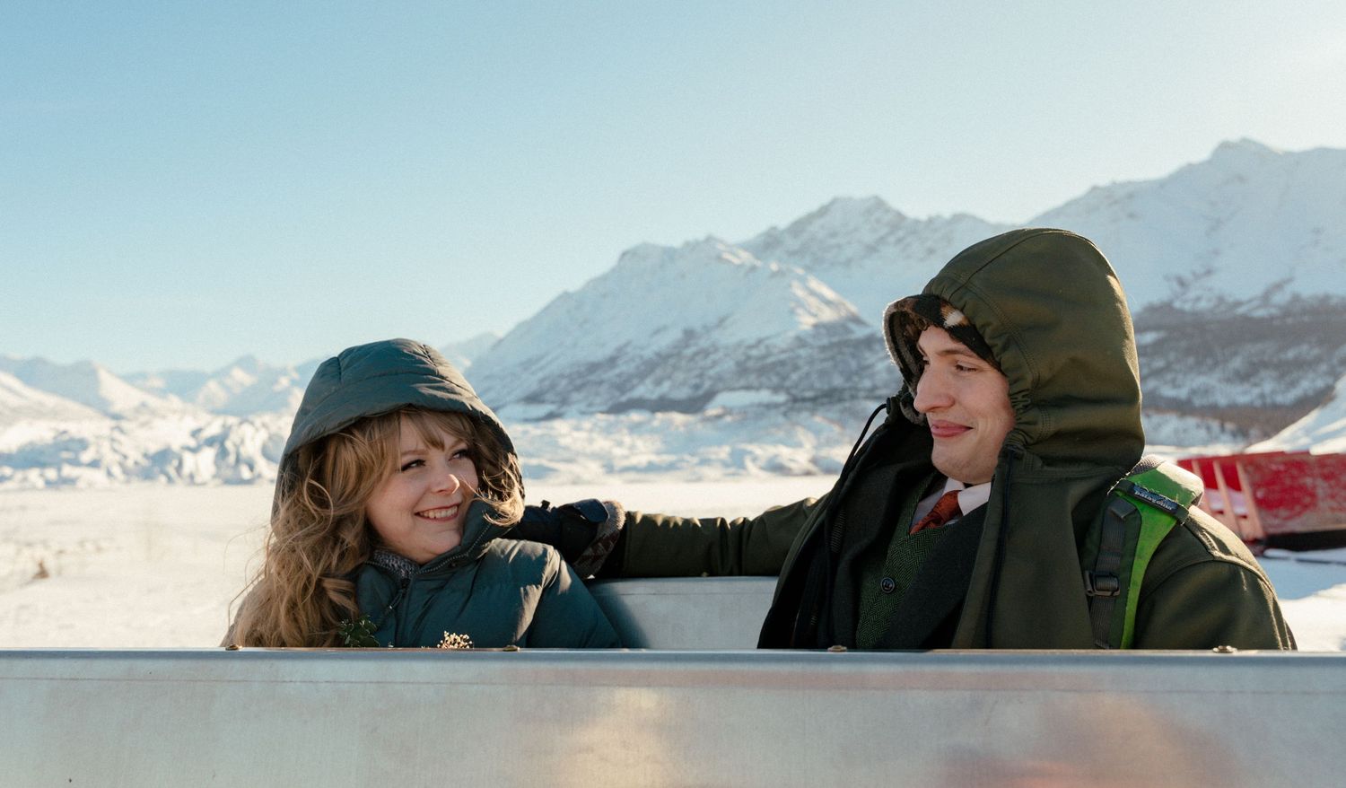 Two people in winter jackets smile at each other with snowy mountain peaks in the background.
