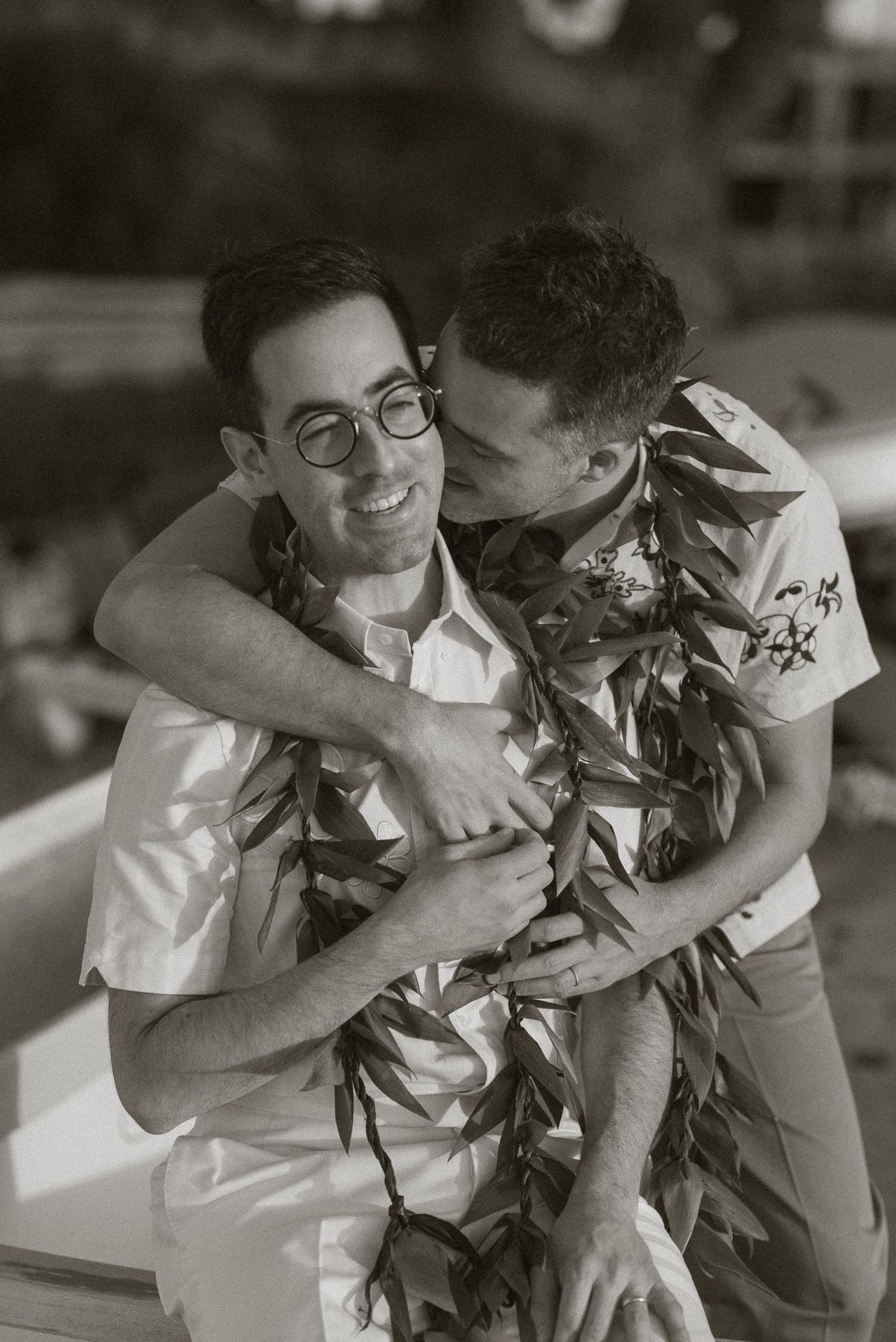 Black and white photo of two grooms embracing joyfully, wearing traditional Hawaiian lei garlands.