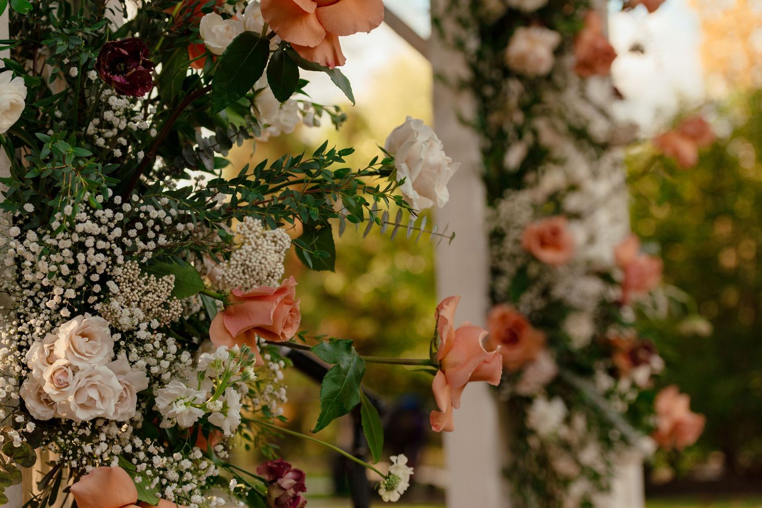 Elegant wedding arch adorned with peach roses, white blooms, baby's breath, and lush greenery in an outdoor ceremony setting.