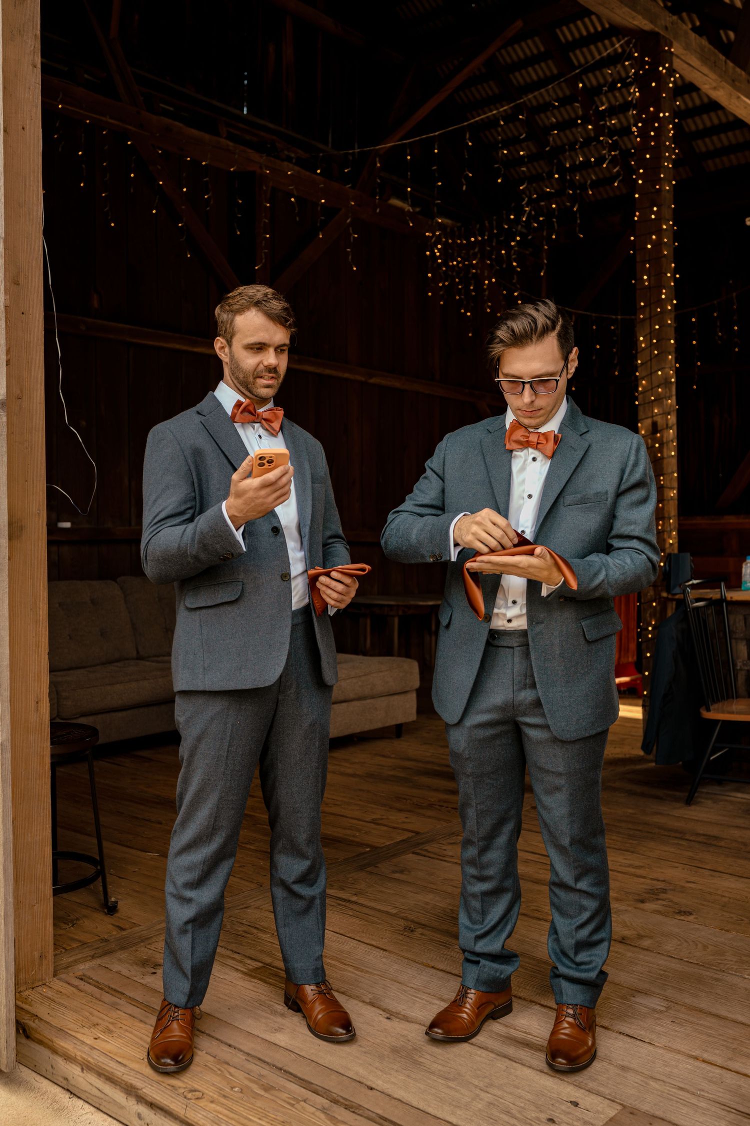 Two groomsmen in matching blue suits and orange bow ties stand in a rustic barn venue decorated with fairy lights.