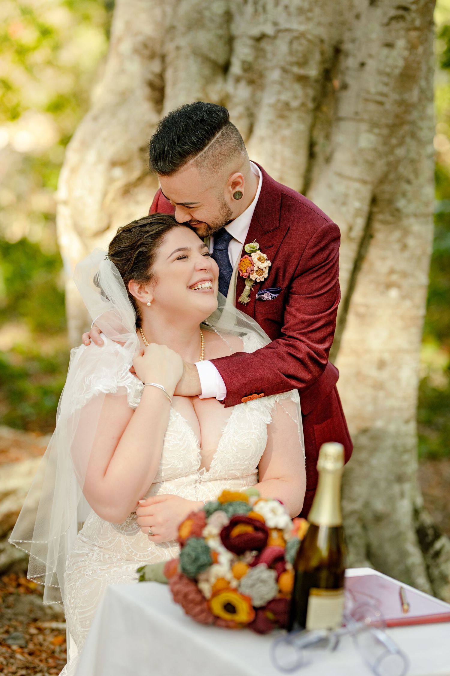 Bride and groom share a tender moment under a tree, with a bouquet and champagne bottle on the table before them.