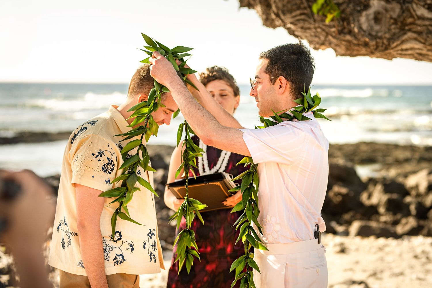 Couple exchanging lei during Hawaiian beach wedding ceremony with officiant and ocean backdrop.