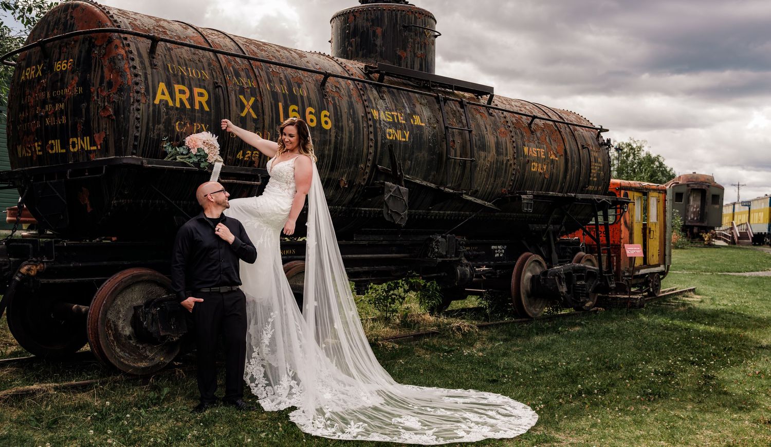Bride and groom pose beside a vintage rusty train tanker car outdoors at a historic railway yard on their wedding day.