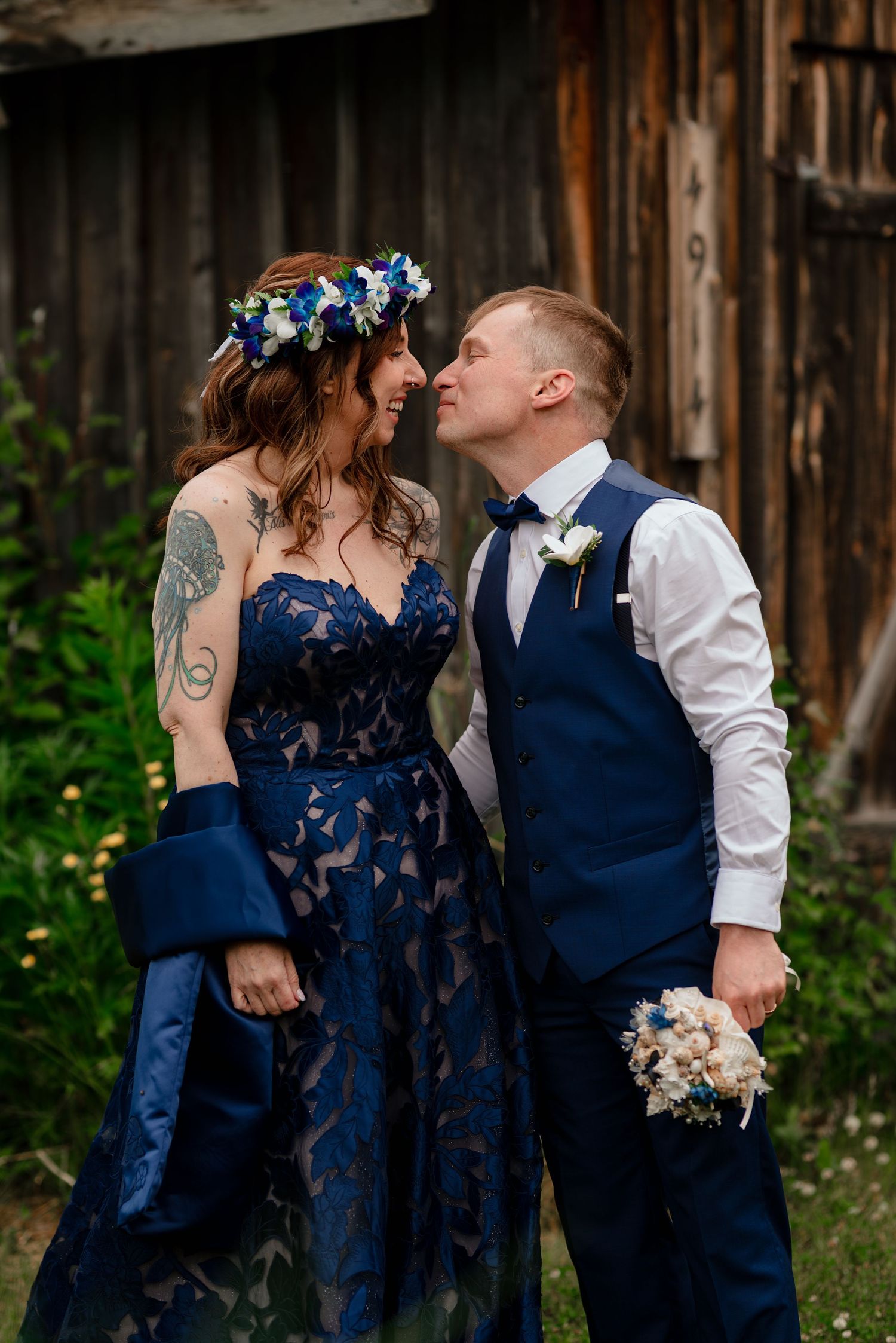 Couple in navy blue wedding attire laughing together outdoors, bride wearing floral crown and lace gown, groom in suit with boutonniere.