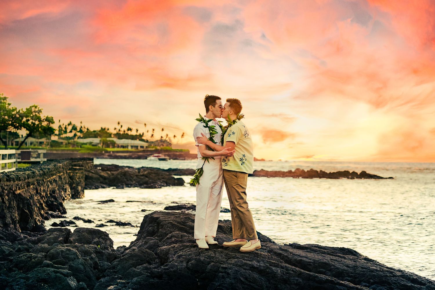 Two grooms kiss on volcanic rocks during a stunning Hawaiian sunset beach wedding ceremony.