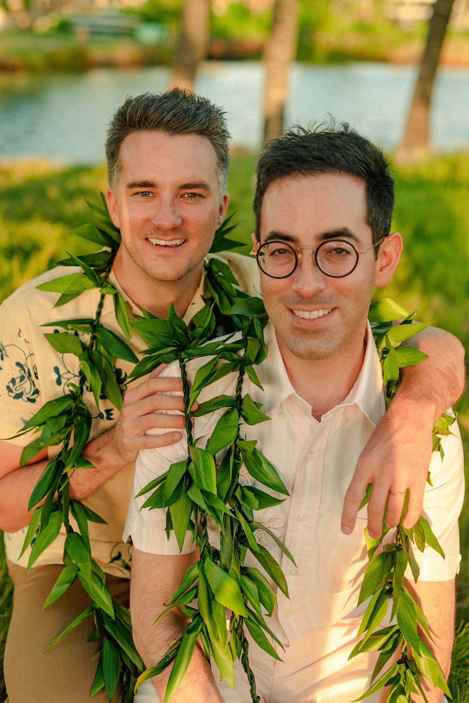 Two smiling men wearing green leaf leis pose together outdoors in a tropical setting during golden hour.