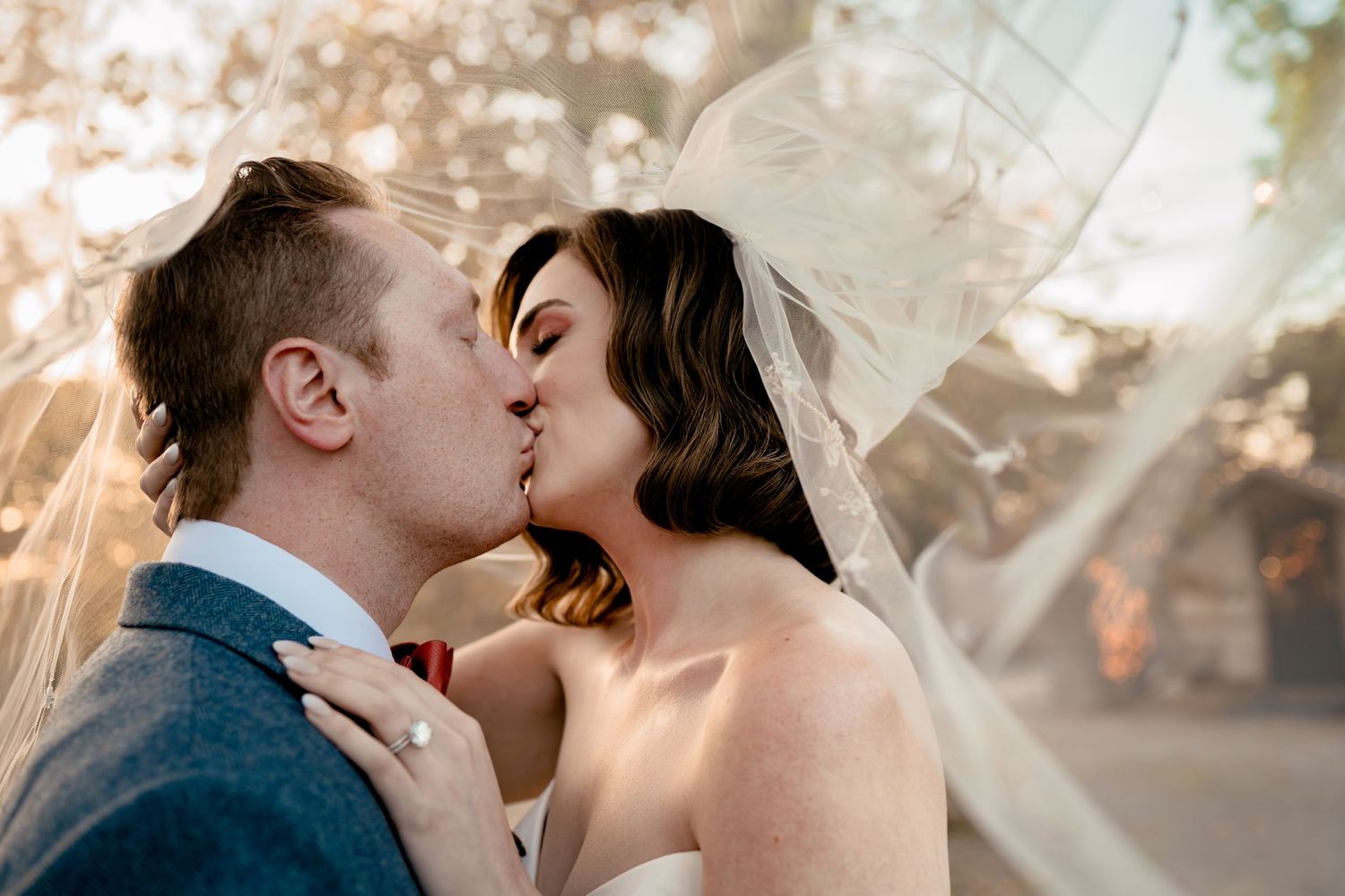 Bride and groom share a kiss under their veil as the light peeks through the trees.