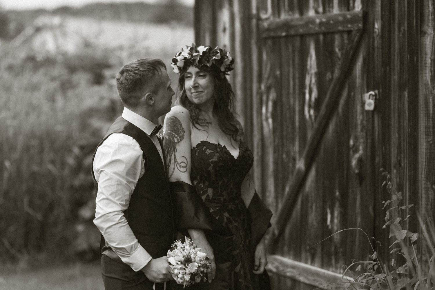Black and white photo of a bride in floral crown and groom embracing by a rustic barn door.
