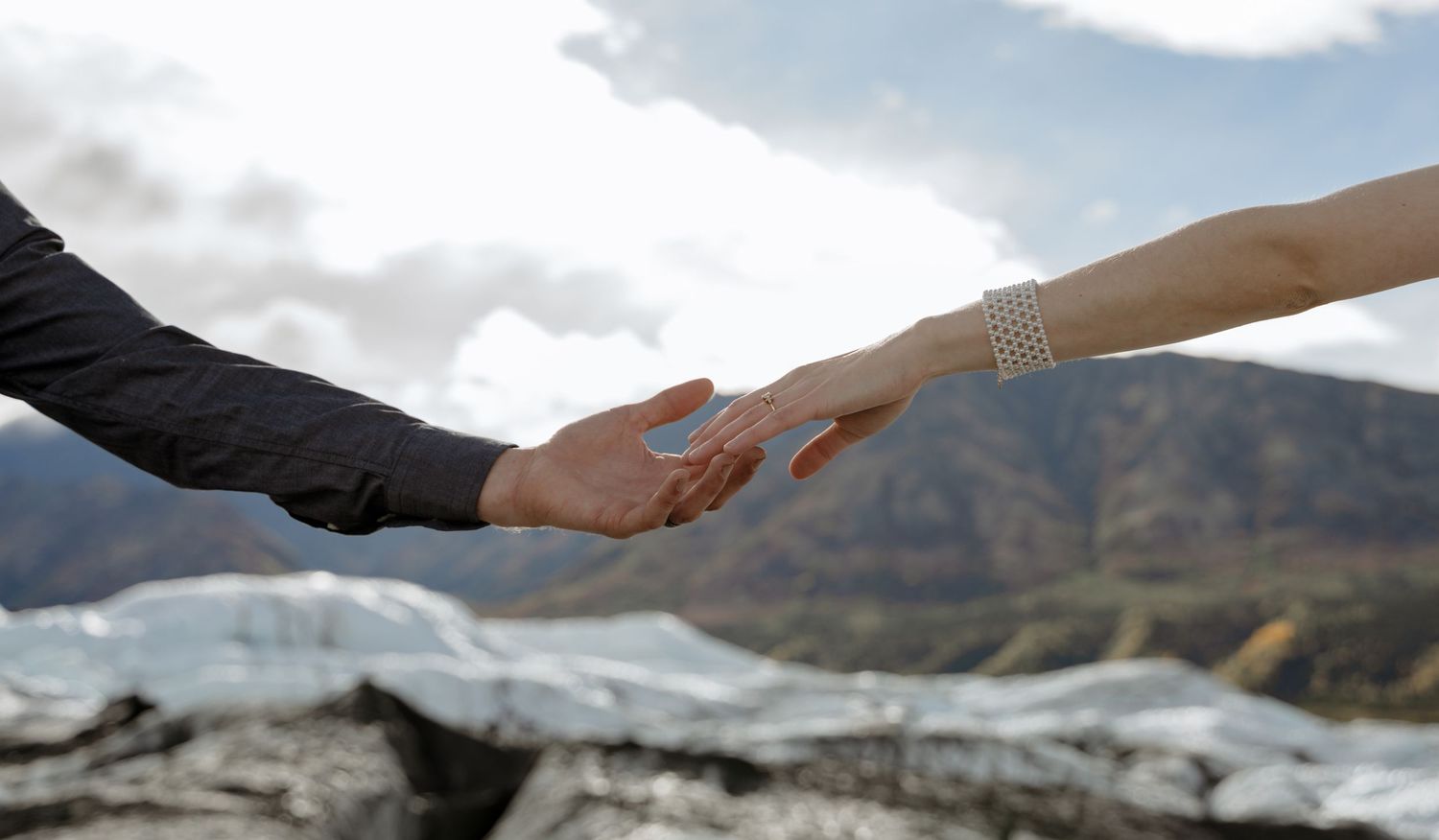 Two hands reaching toward each other against a mountain backdrop, one adorned with a pearl bracelet.