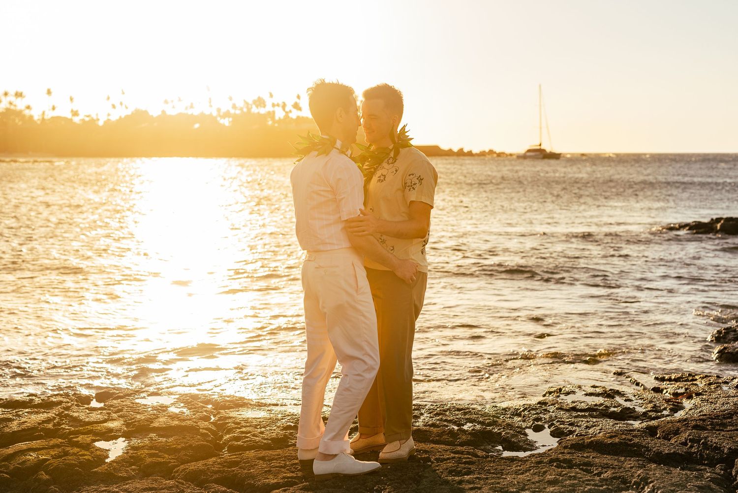 Two grooms embracing romantically on a rocky Hawaiian beach during a breathtaking golden sunset.