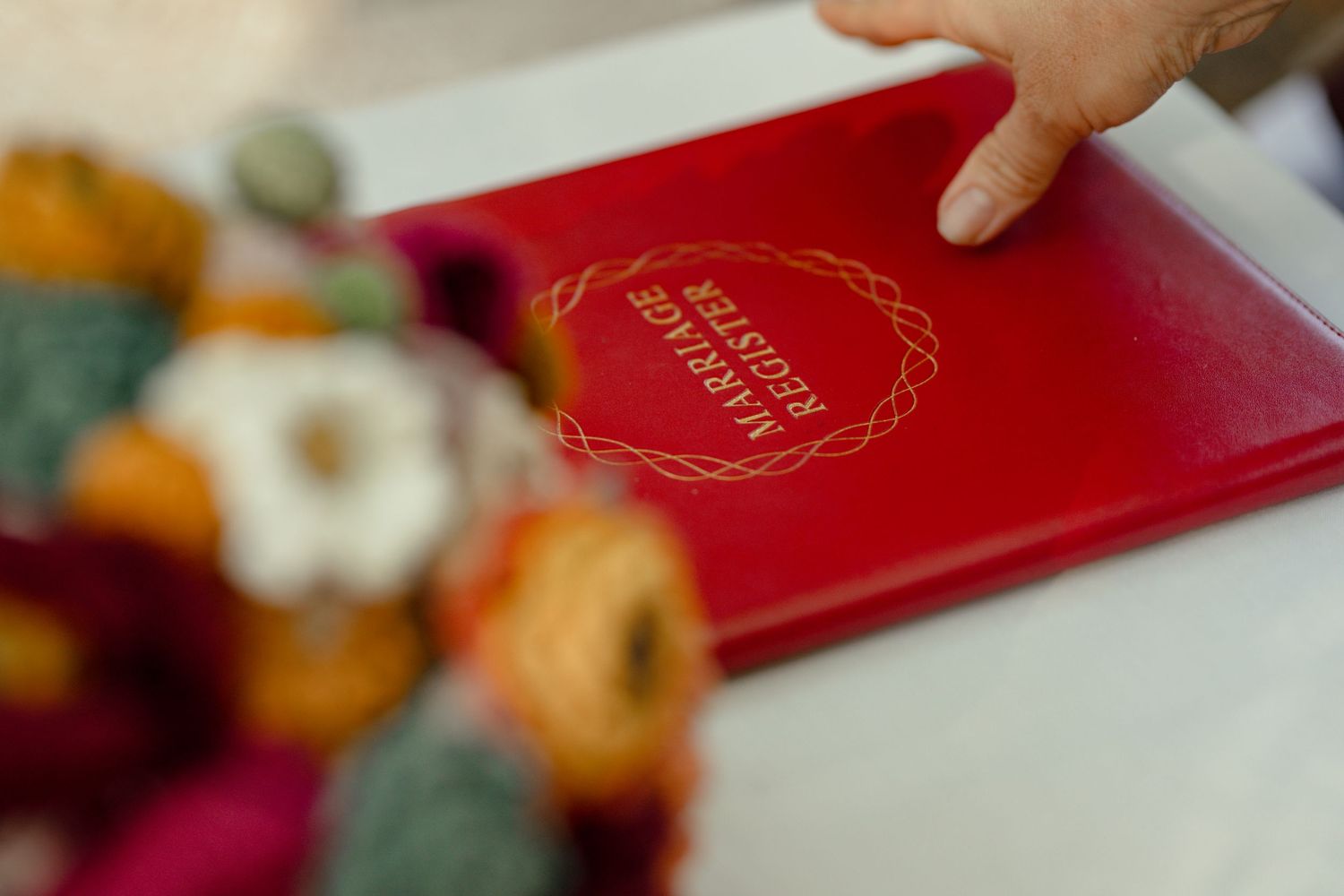 Close-up of red marriage register book with gold lettering beside colorful wedding bouquet on white table.
