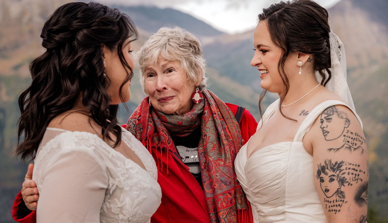 Two brides in white dresses share a tender moment with an elderly woman in red at an outdoor mountain wedding.