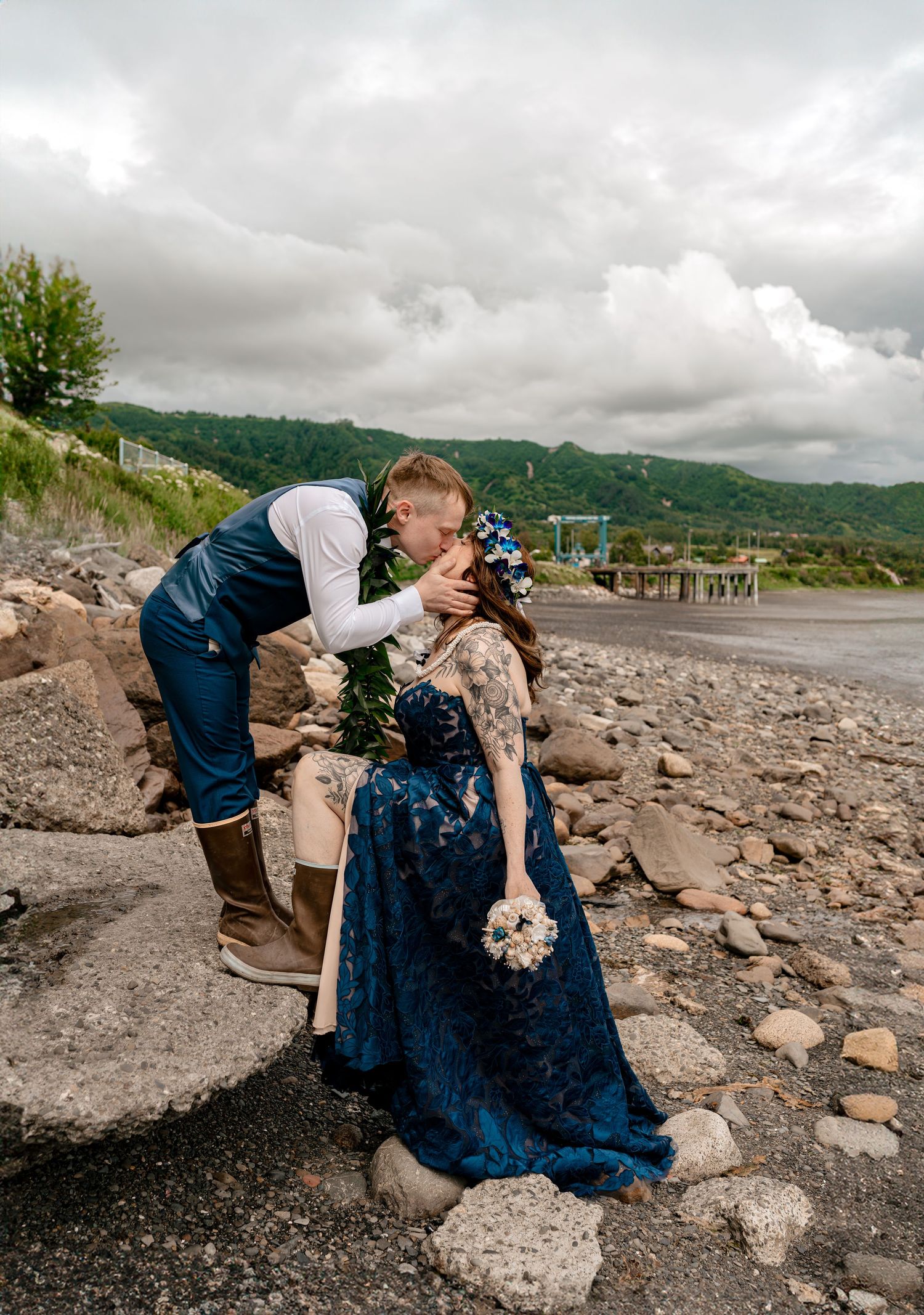 A couple shares a romantic kiss on a rocky shoreline in Homer Alaska. the bride in a navy blue gown with floral crown, shell bouquet.