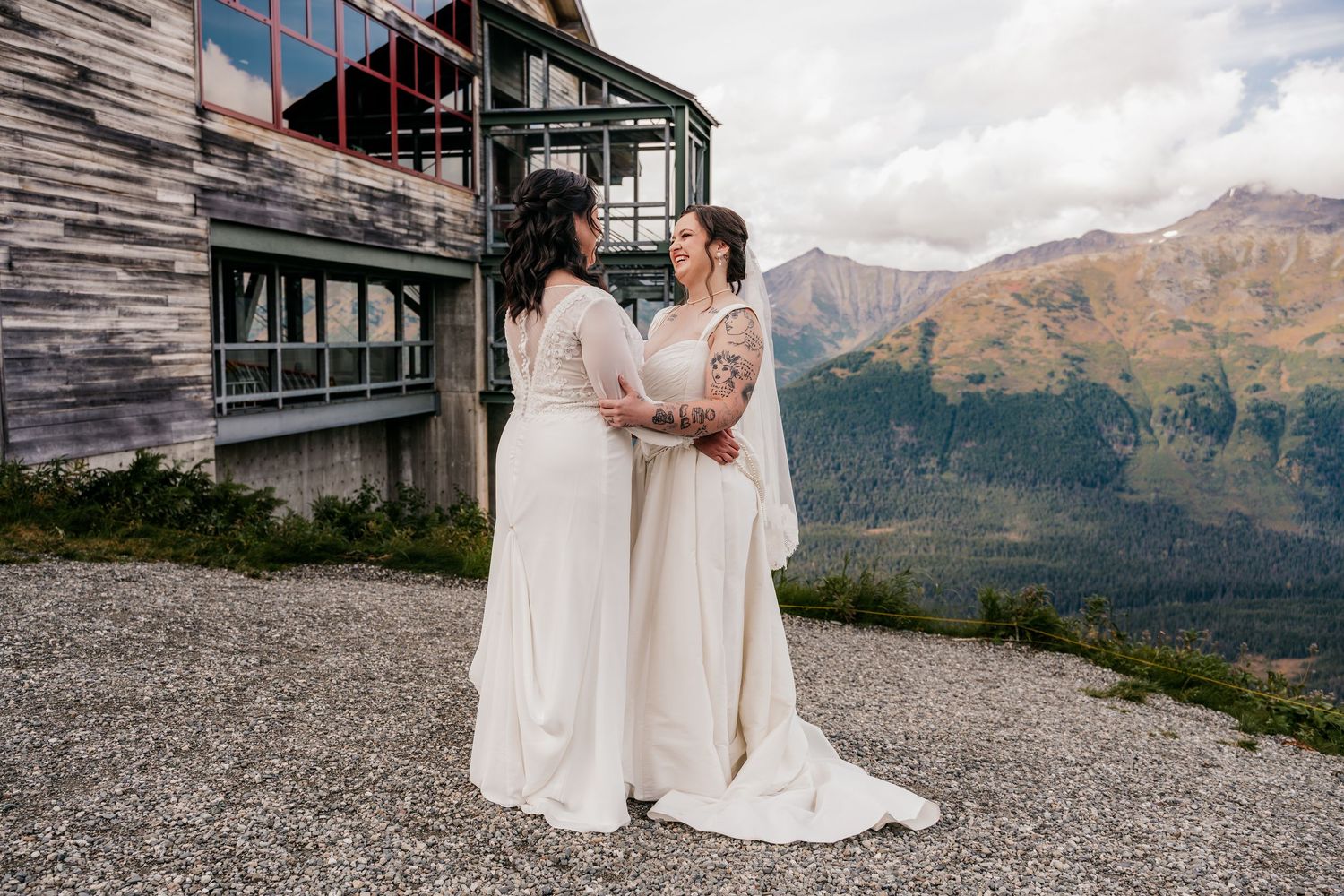Two brides in white gowns hold hands at a mountain wedding venue with scenic alpine views.