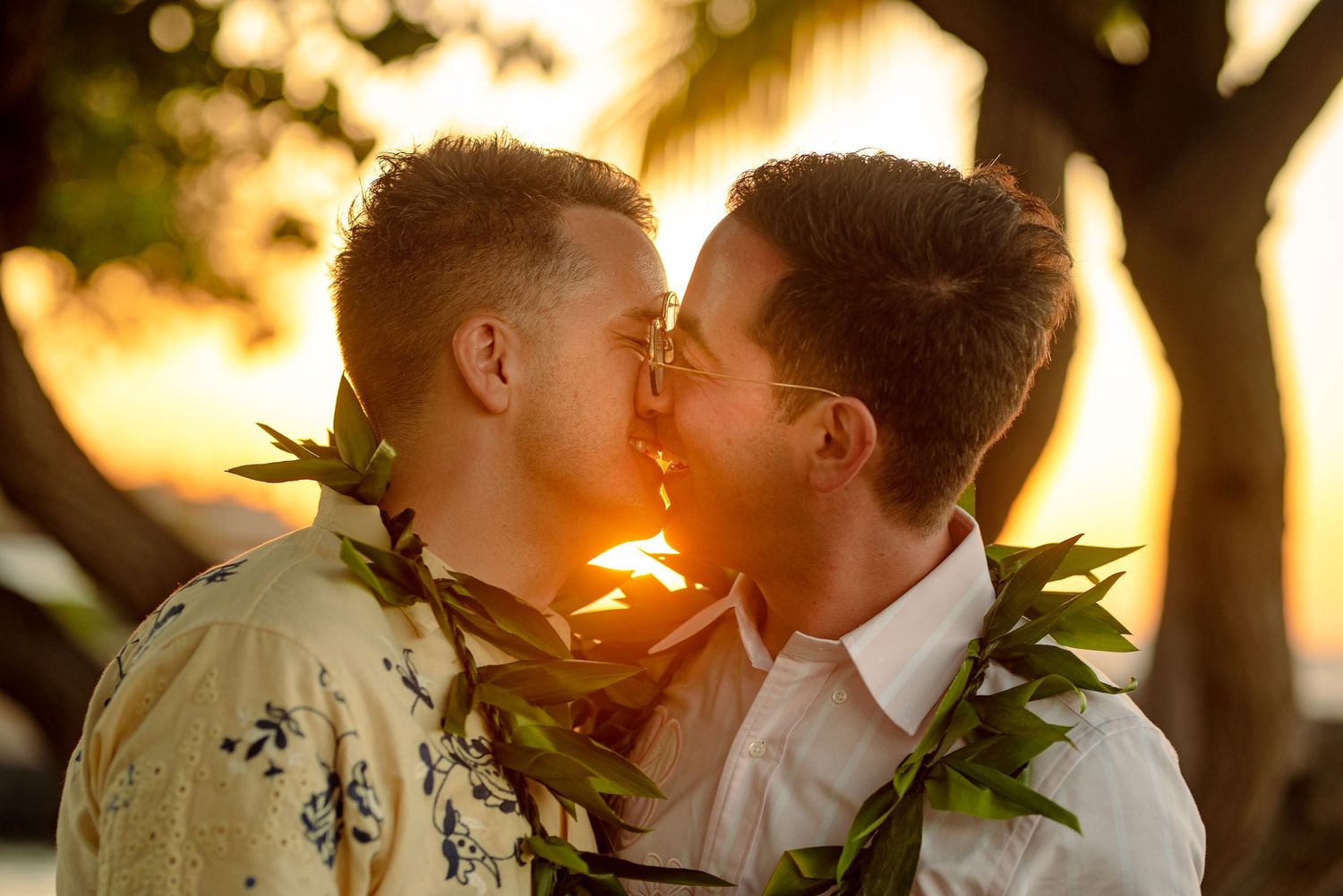 Two men sharing a romantic kiss at sunset while wearing traditional Hawaiian leaf leis.