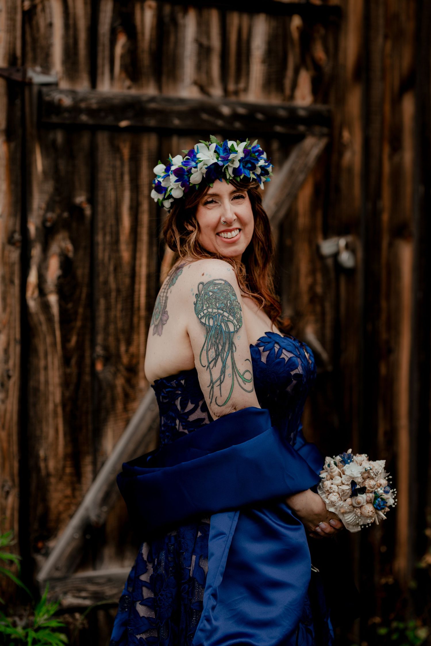 Smiling bride in blue strapless gown with floral crown and jellyfish tattoo holds bouquet before rustic wooden barn doors.