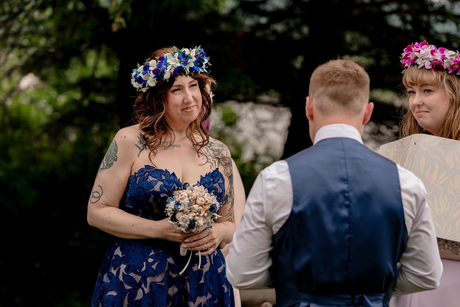 Bride in blue floral dress and flower crown holds bouquet at outdoor wedding ceremony.
