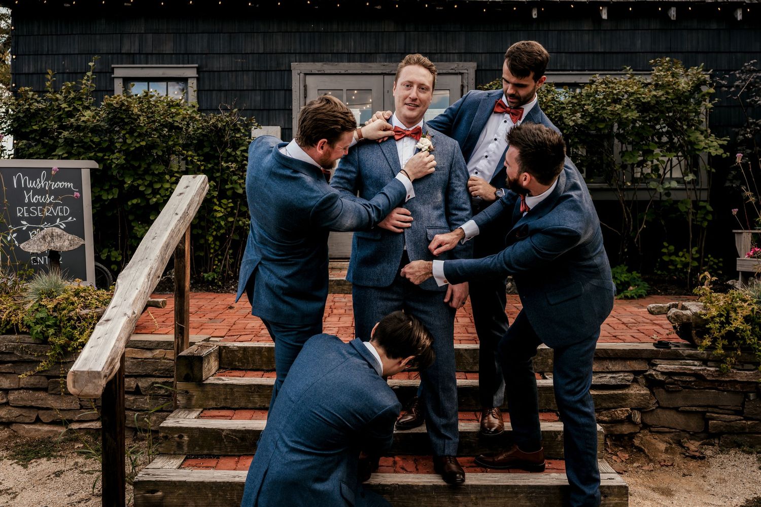 Groomsmen in navy suits playfully helping the groom get ready on rustic wooden steps at an outdoor wedding venue.