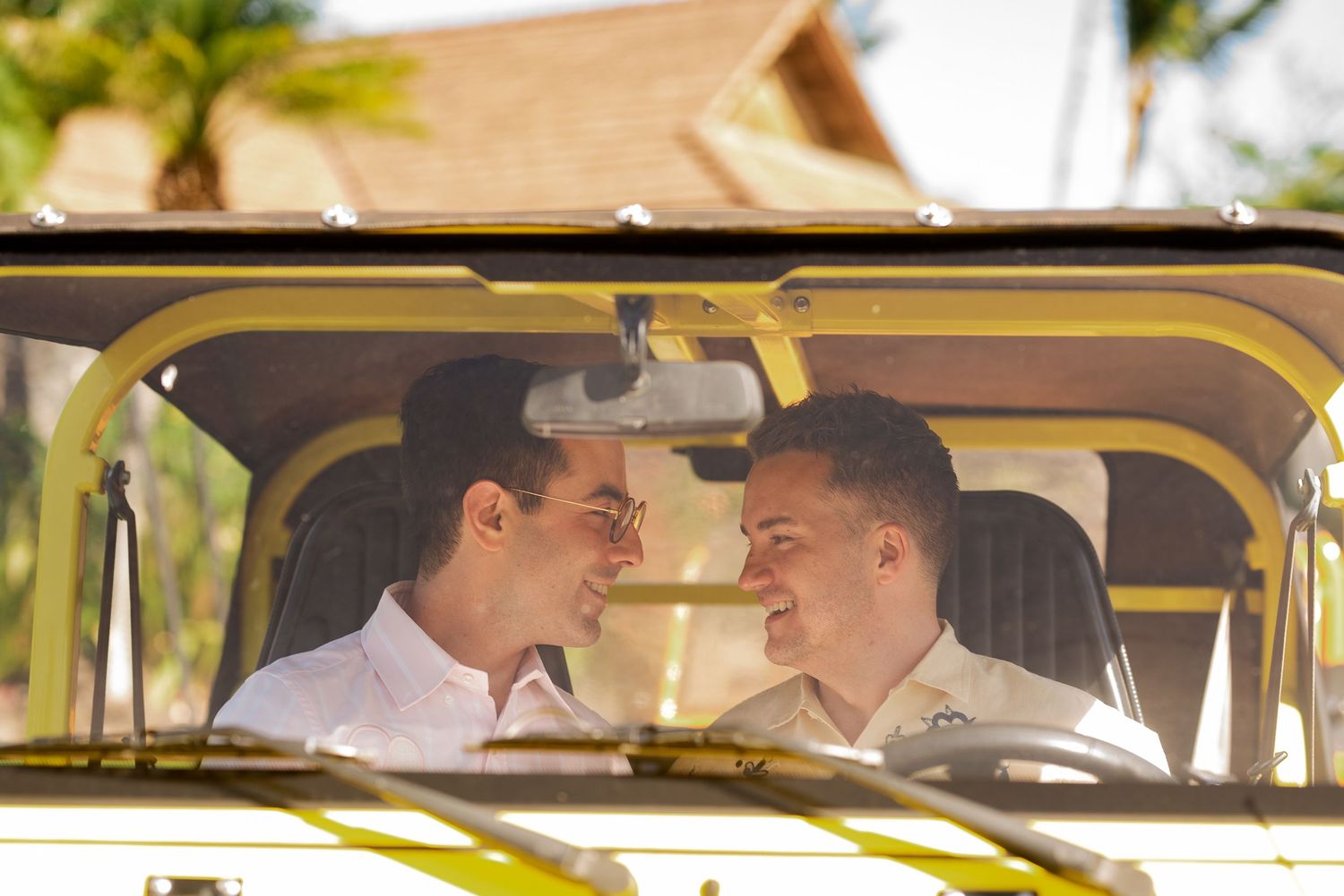 Two men sharing a romantic moment in a yellow open-top jeep surrounded by tropical scenery.