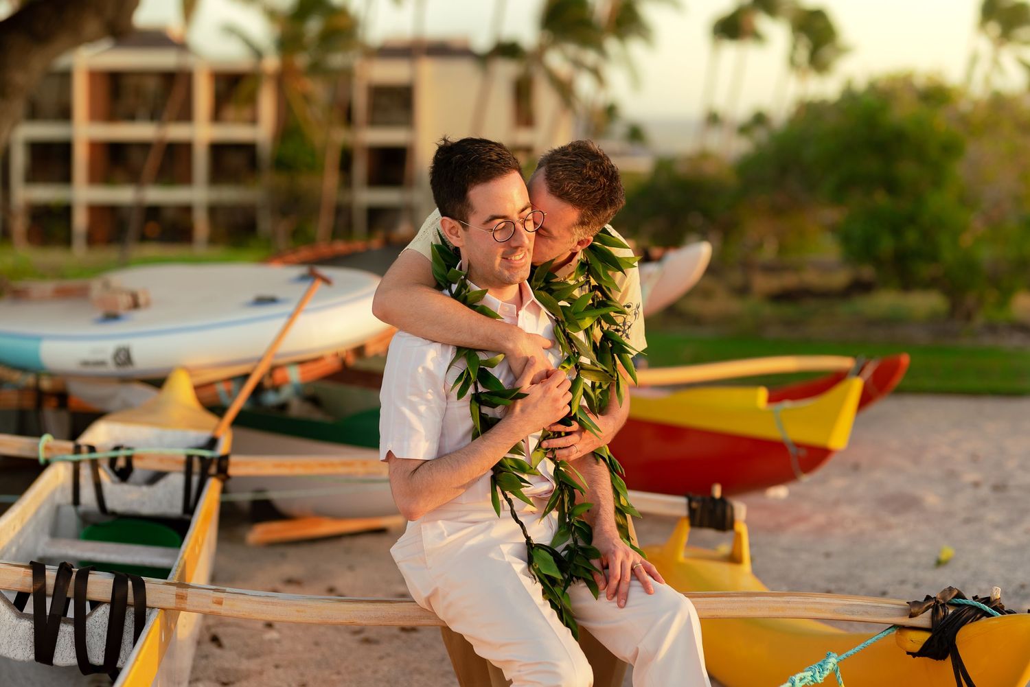 Two men sharing a romantic embrace on a Hawaiian beach with colorful outrigger canoes at sunset.