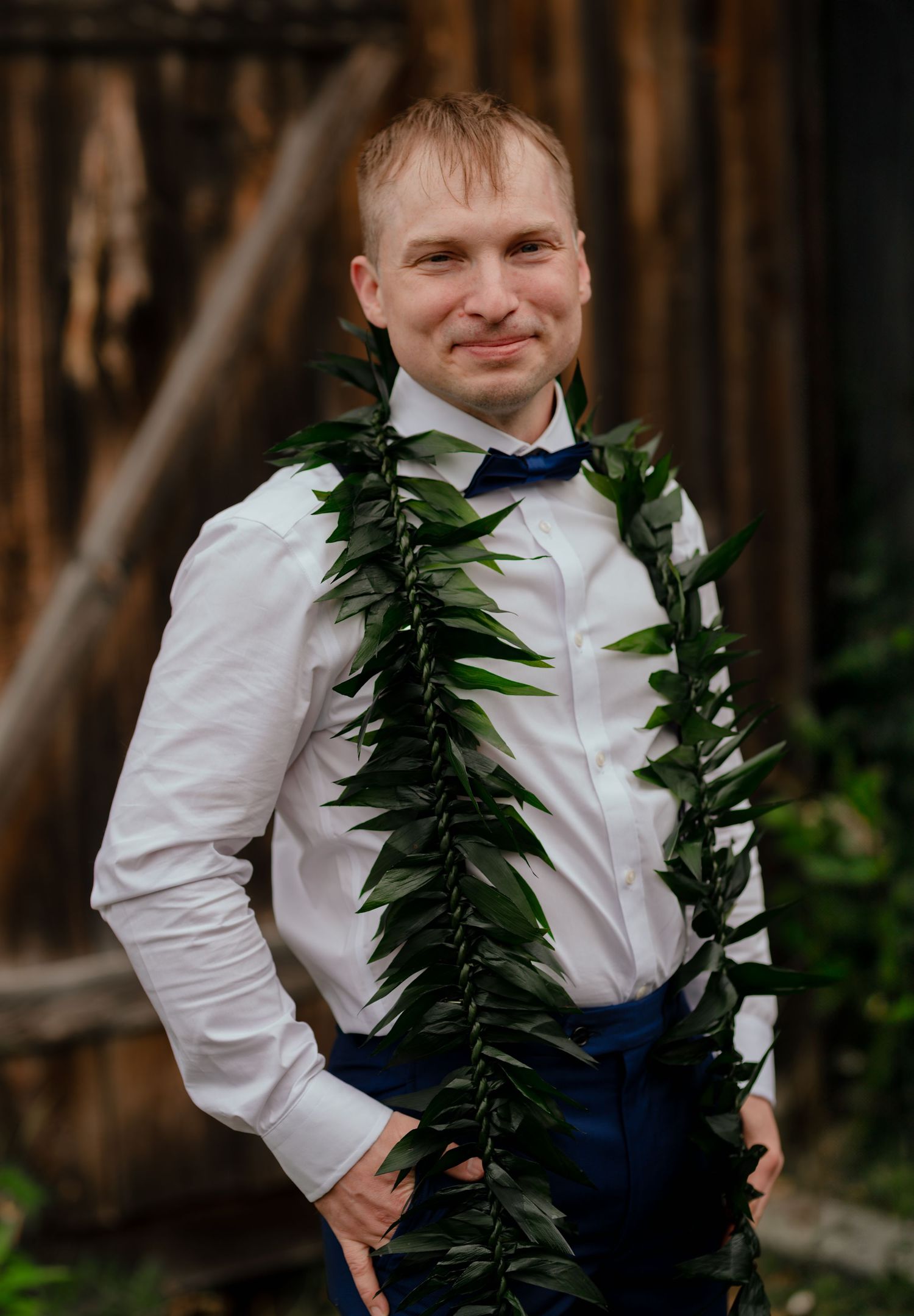Man in white shirt and navy bow tie wearing a green leaf lei, smiling outdoors near rustic wooden backdrop.