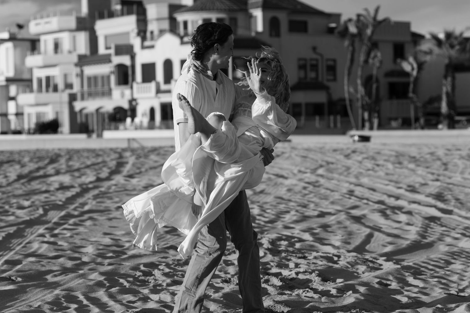 Black and white photo of a couple embracing on a sandy beach with Mediterranean-style buildings in the background.