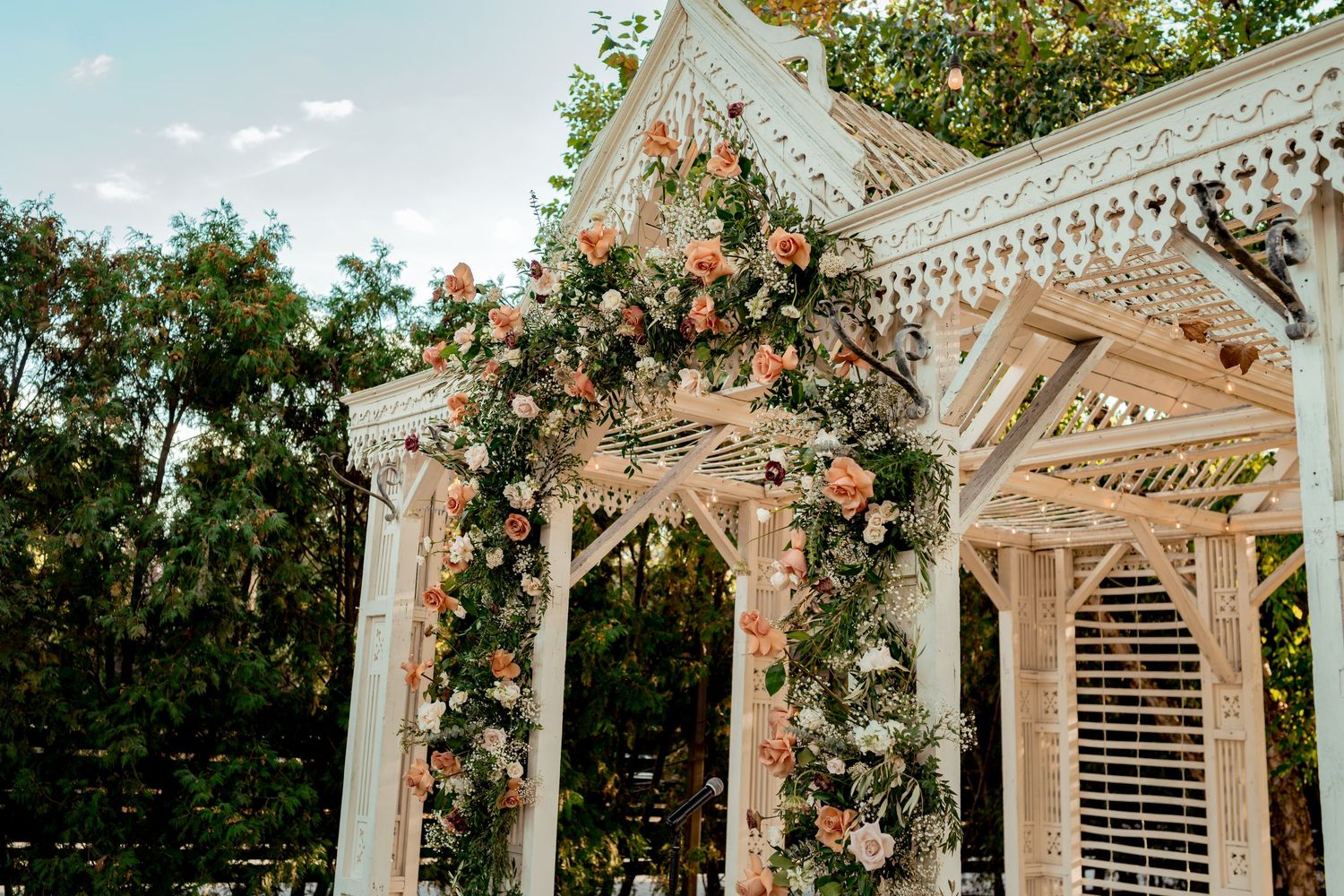 White gazebo adorned with lush peach and pink floral arrangements and string lights for an elegant outdoor wedding.