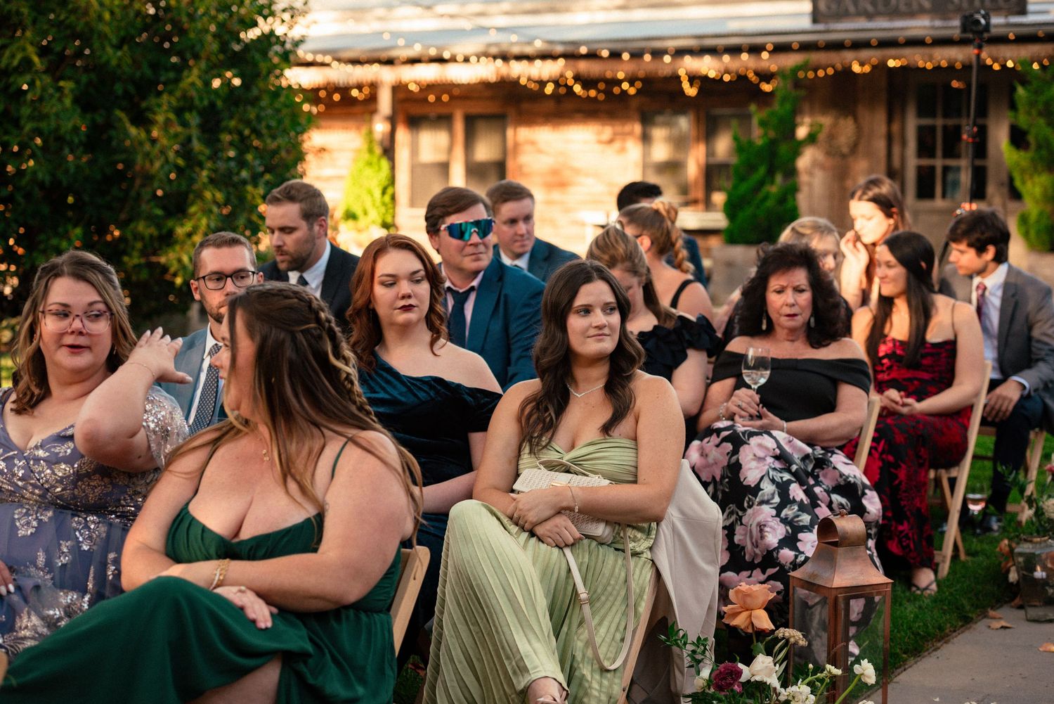 Guests in formal attire seated outdoors at an evening wedding ceremony, surrounded by lush greenery and warm string lights.