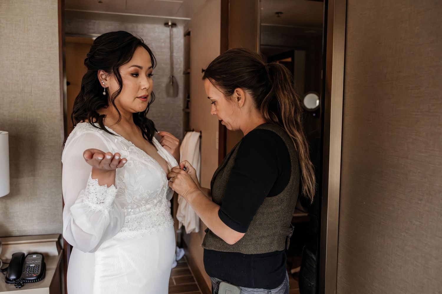 A bride in a white wedding gown gets help fastening her dress from a woman in a gray vest inside a hotel room.