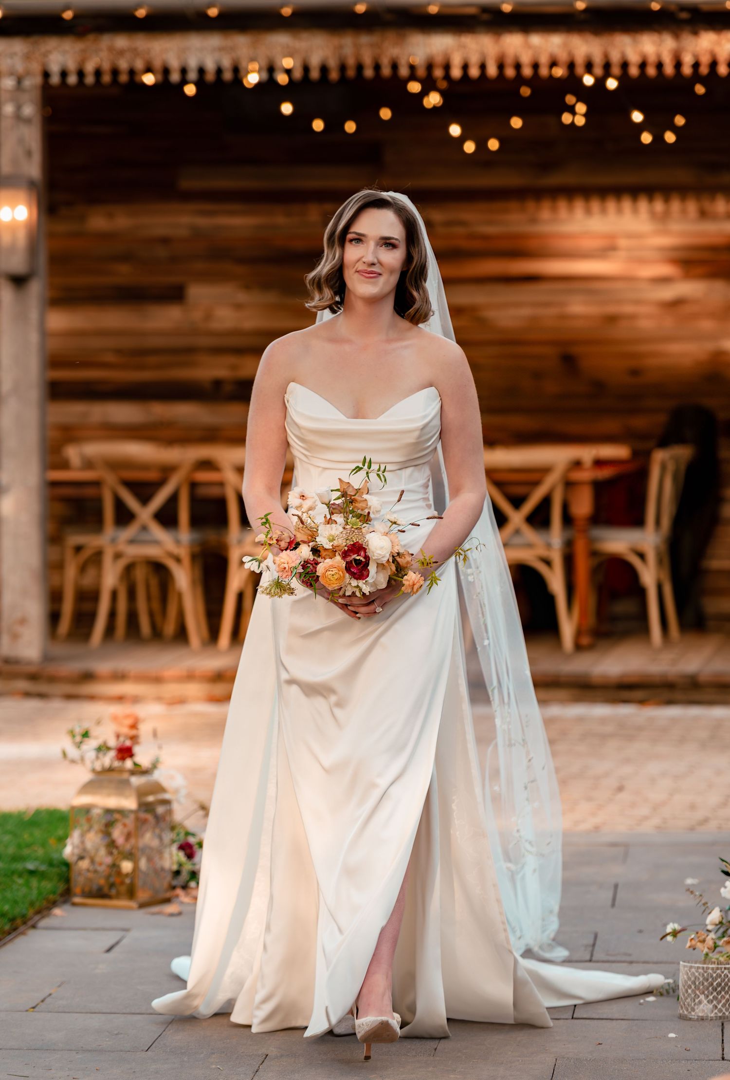 Bride in strapless ivory gown with veil holds floral bouquet, standing before rustic barn with string lights.