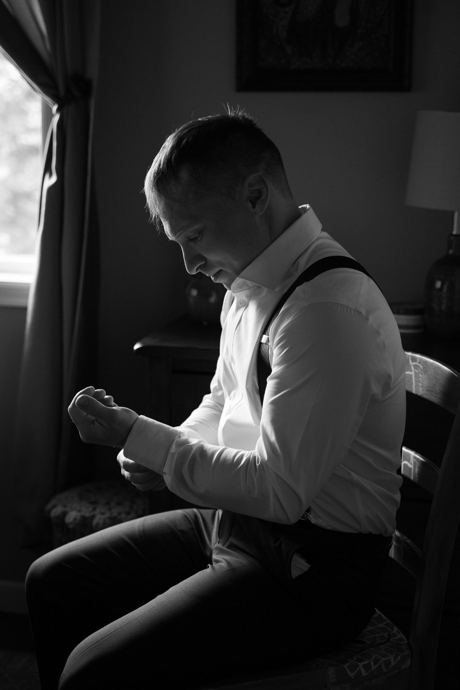Black and white photo of a groom in suspenders fastening his cufflinks while seated by a window.