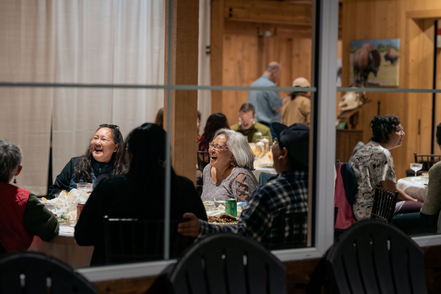 Group of people laughing and dining together at a rustic indoor gathering, enjoying a warm social meal.