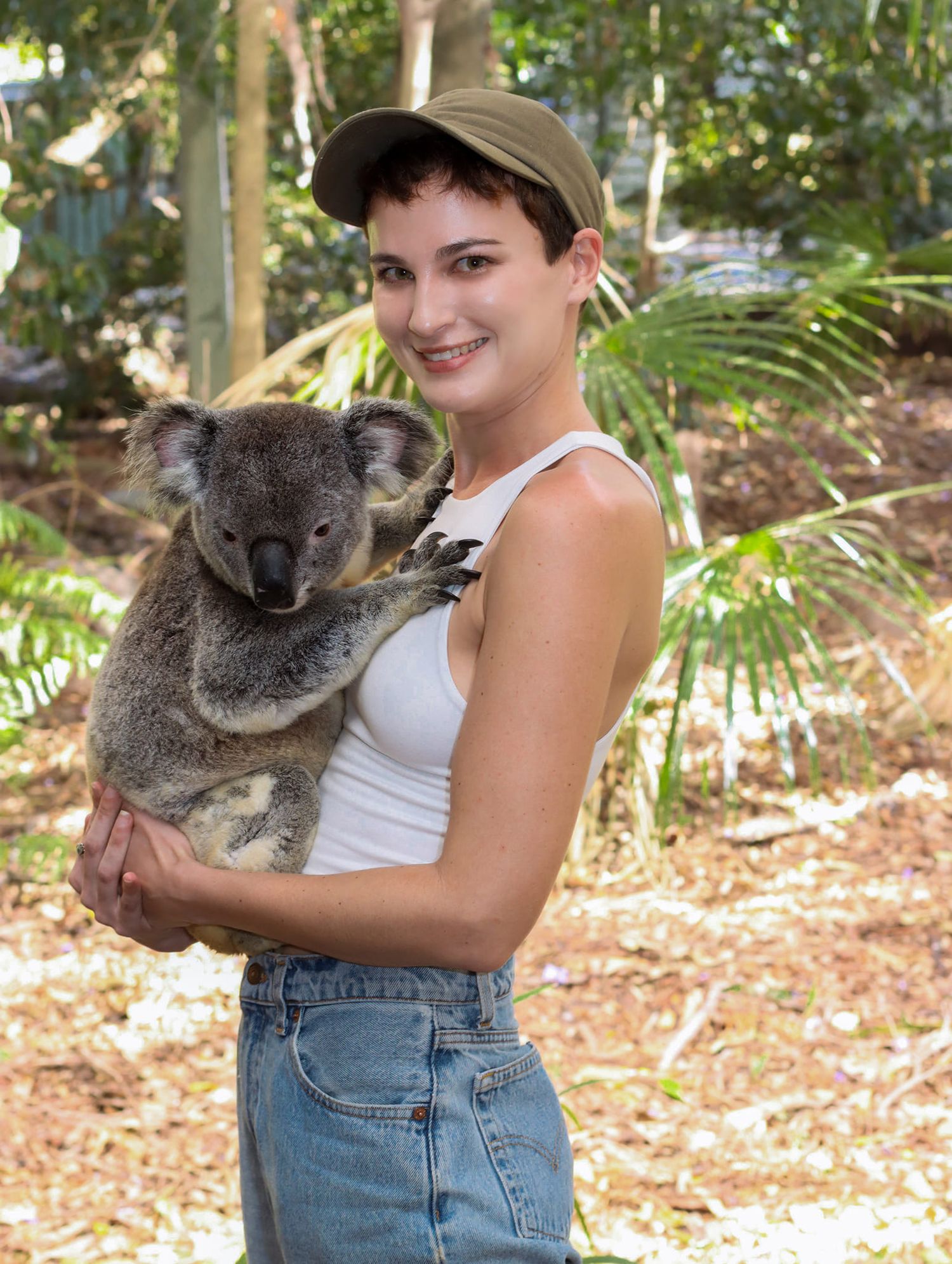 A smiling young woman in a cap and jeans holds a koala bear outdoors in a lush Australian wildlife park.