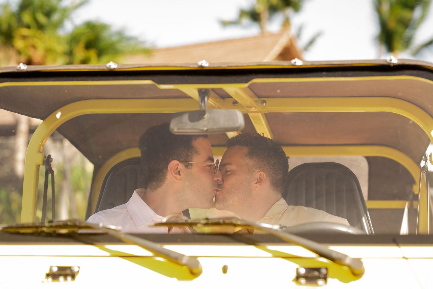 Two grooms sharing a romantic kiss inside a vintage yellow vehicle on their wedding day.