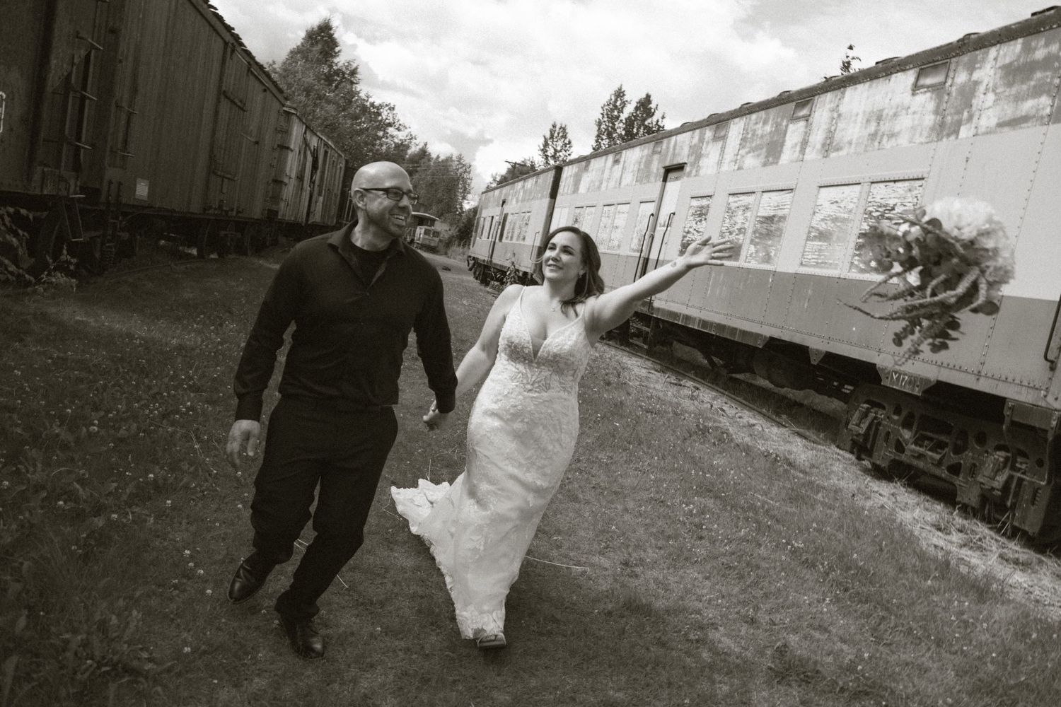 Black and white photo of bride and groom walking hand in hand along railroad tracks with vintage train cars in background.