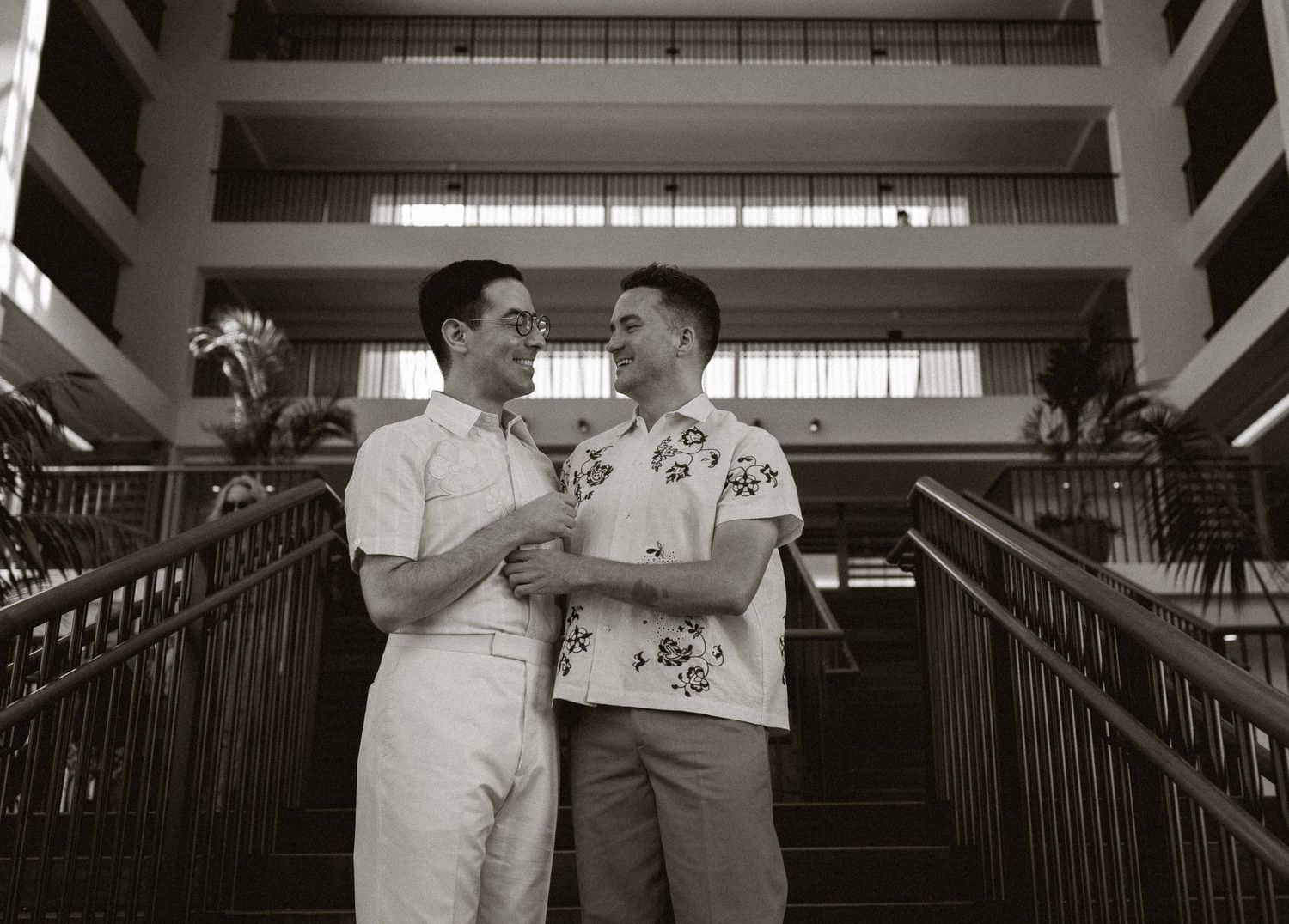 Two men smiling and holding hands on a staircase in a modern building, sharing a tender moment.
