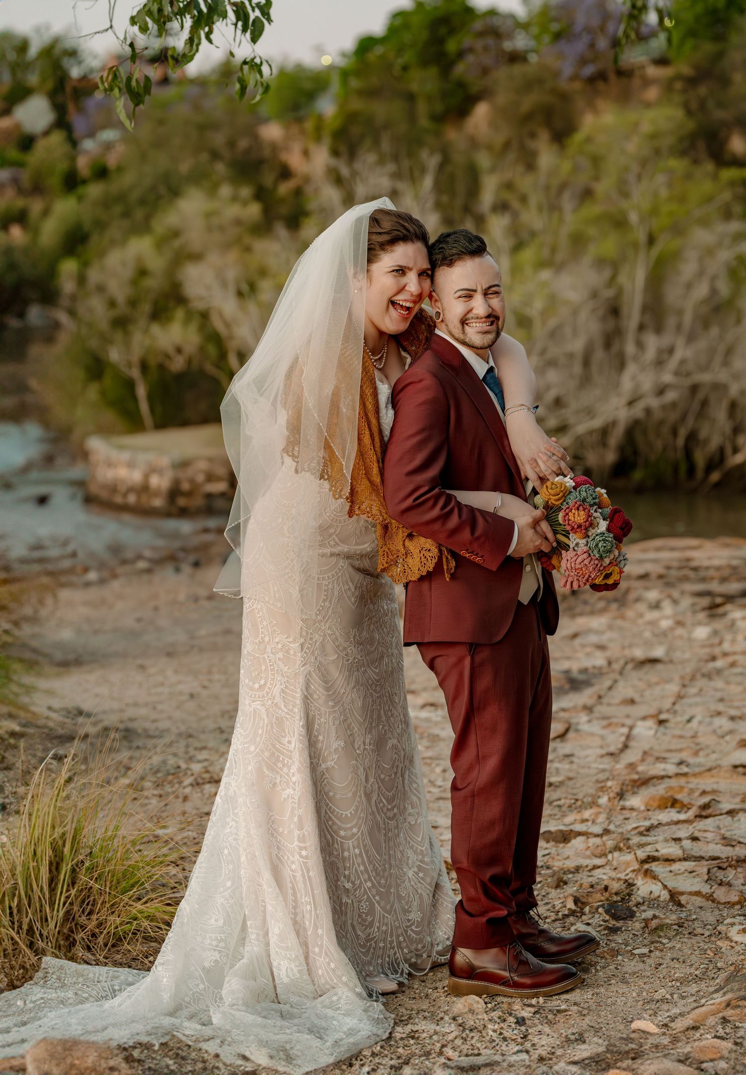 Laughing bride in lace gown and veil poses back-to-back with groom in burgundy suit holding colorful bouquet outdoors.