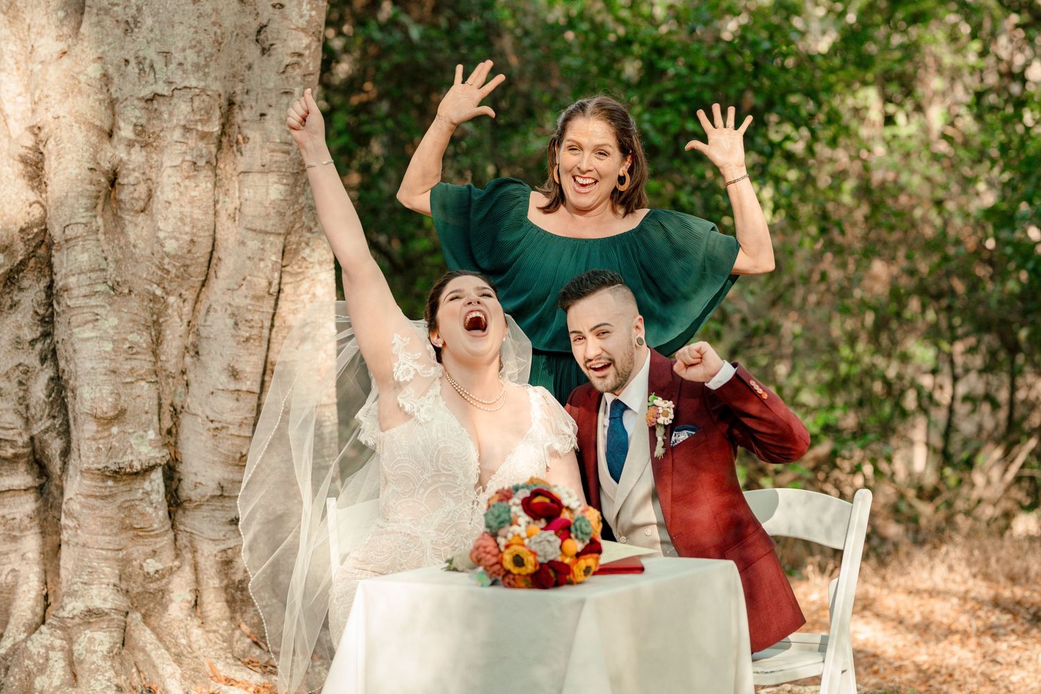 Joyful bride and groom celebrate at outdoor wedding table with excited officiant raising hands behind large tree.