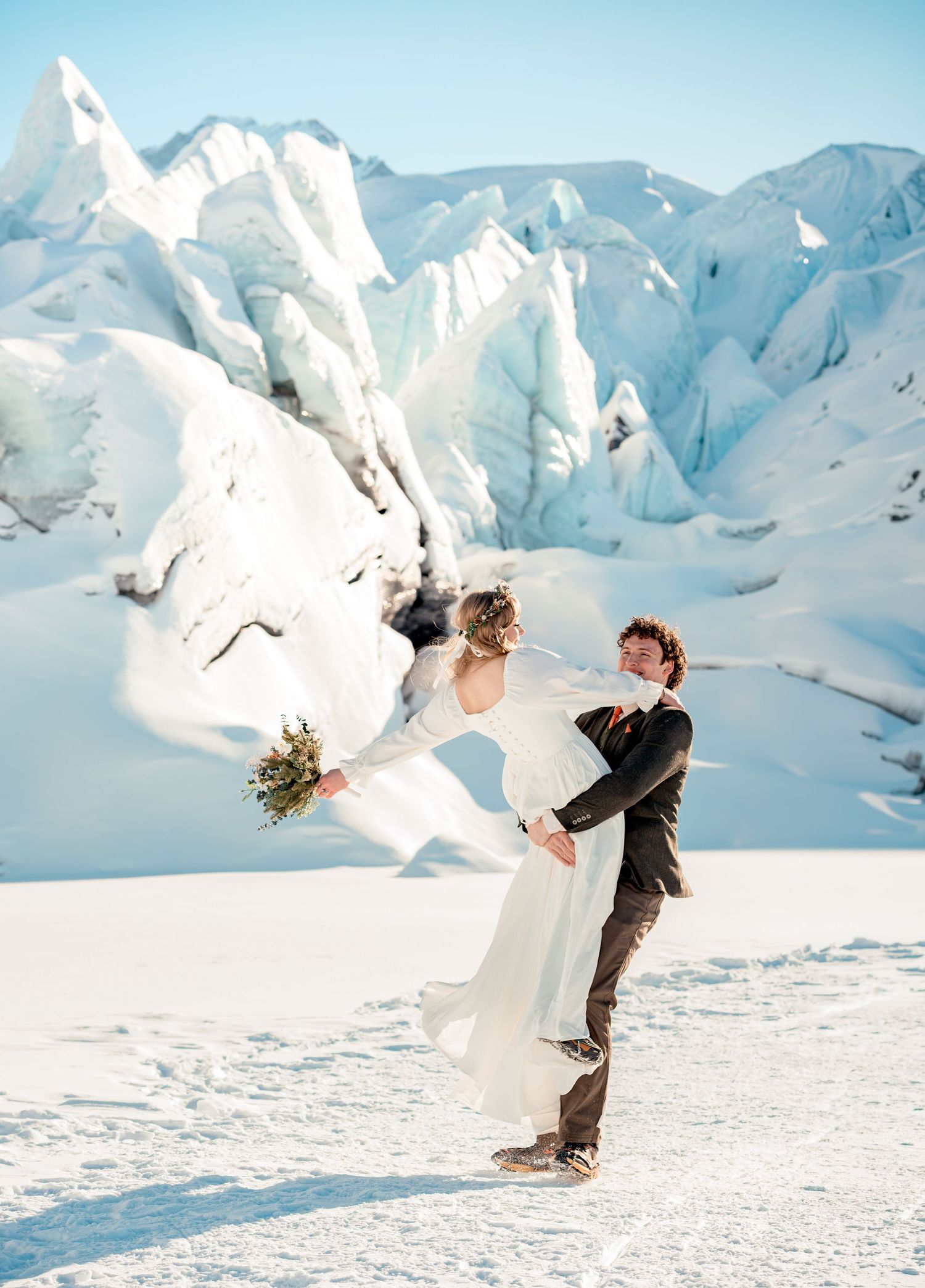 Groom lifting bride in white gown on snowy glacier landscape with dramatic ice formations during winter wedding photoshoot.