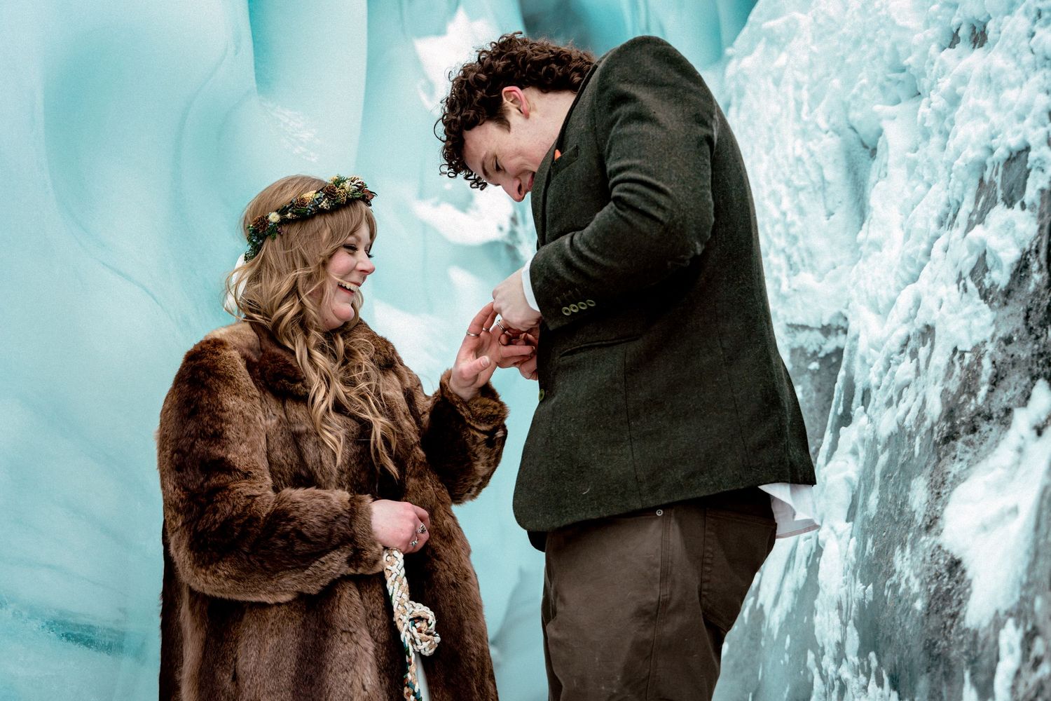 Couple exchanging rings in a stunning ice cave, bride wearing floral crown and fur coat during winter wedding ceremony.