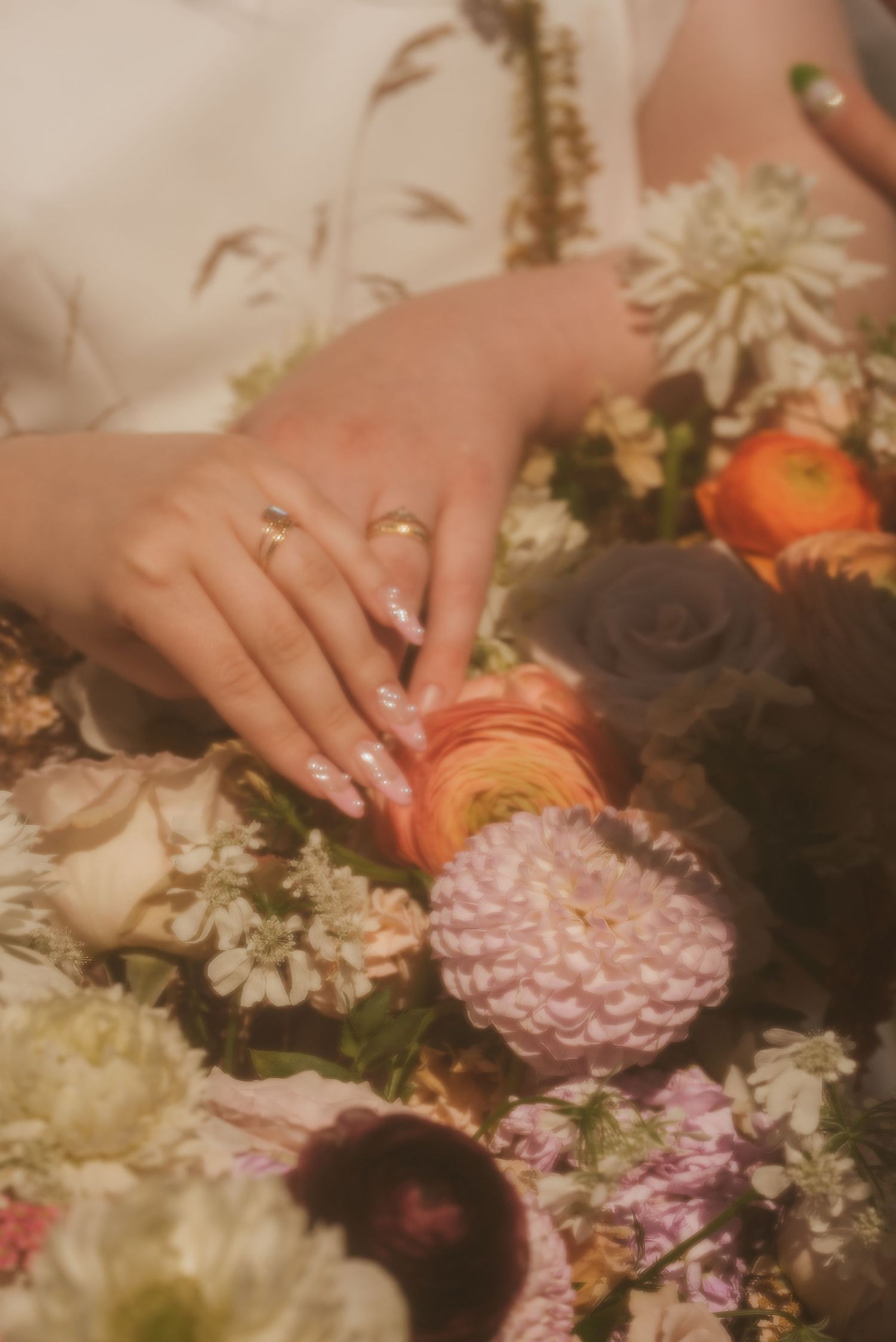 Elegant hands with gold rings and manicured nails resting on a lush floral arrangement with dahlias and roses.